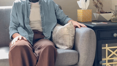 Person sitting on a gray sofa with a beige pillow, wearing a blue jacket and brown pants, next to a dark blue side table with a gold geometric design, a white tissue box, and decorative items.