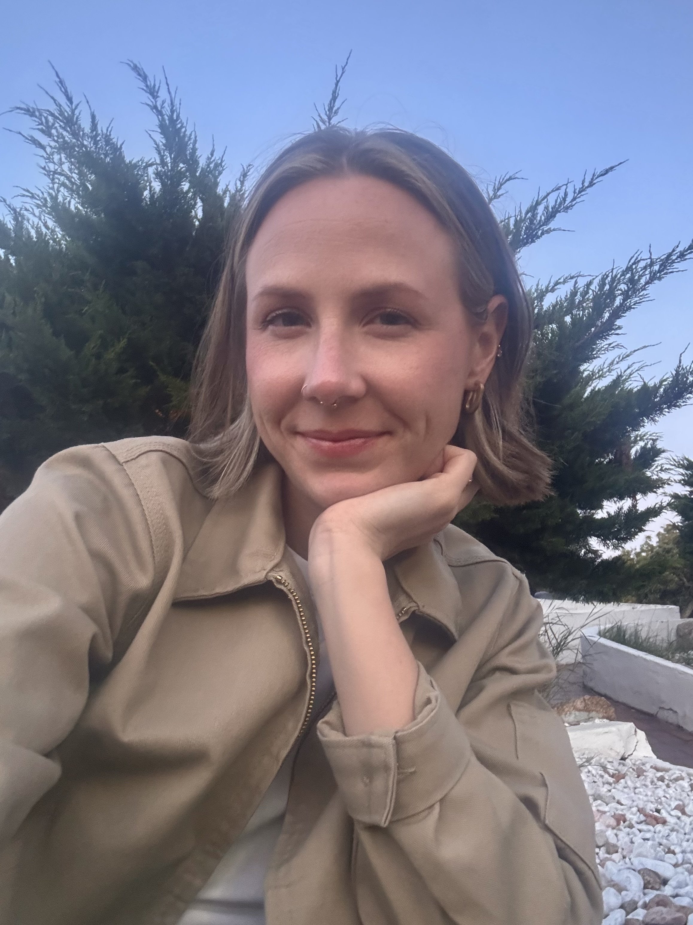 A woman with short hair, wearing earrings and a nose ring, sitting outdoors during winter, smiling slightly, with her chin resting on her hand near her face. There are evergreen trees and snow on the ground in the background.
