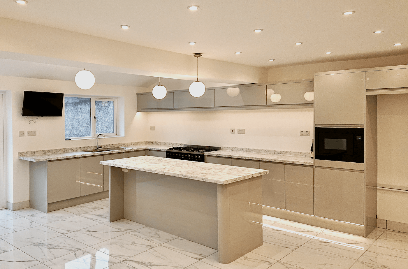 Modern luxury kitchen in Henleaze, Bristol with beige cabinets, marble countertops, an island, a wall-mounted TV, a window with frosted glass, and a microwave built into the cabinetry.