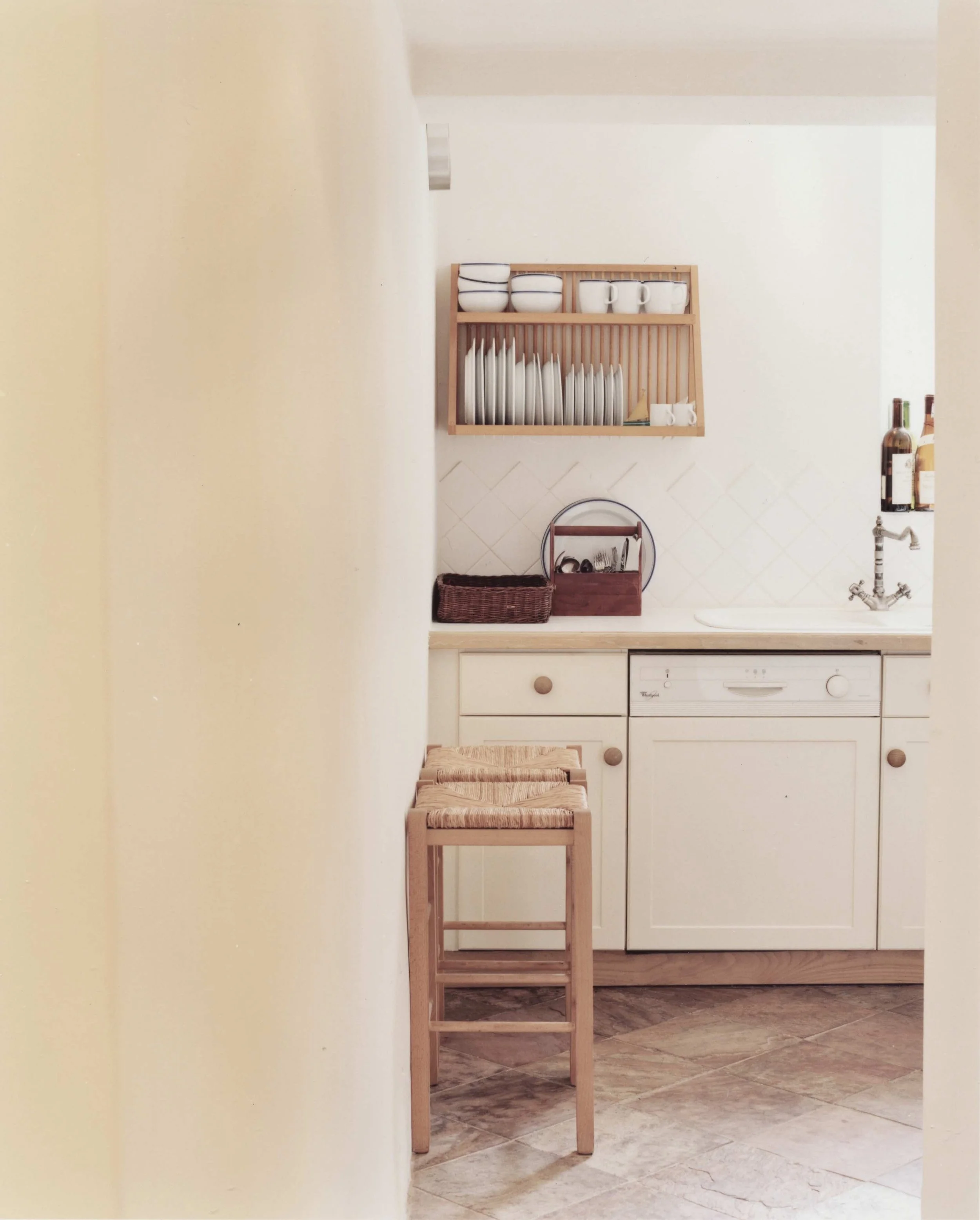 The entrance to a kitchen in Clifton, Bristol with white cabinets, a dish rack with plates, cups, and bowls, a small wooden countertop, a woven basket, and a wooden stool.