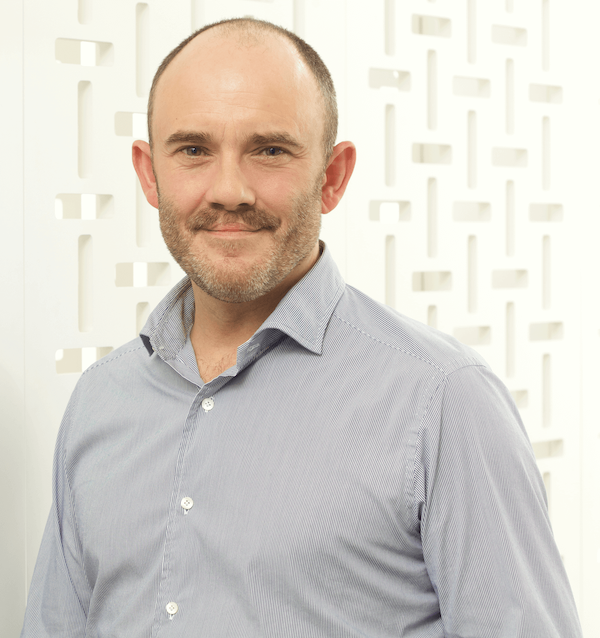 Portrait of a man, wearing a light blue button-up shirt, standing in front of a white decorative wall.