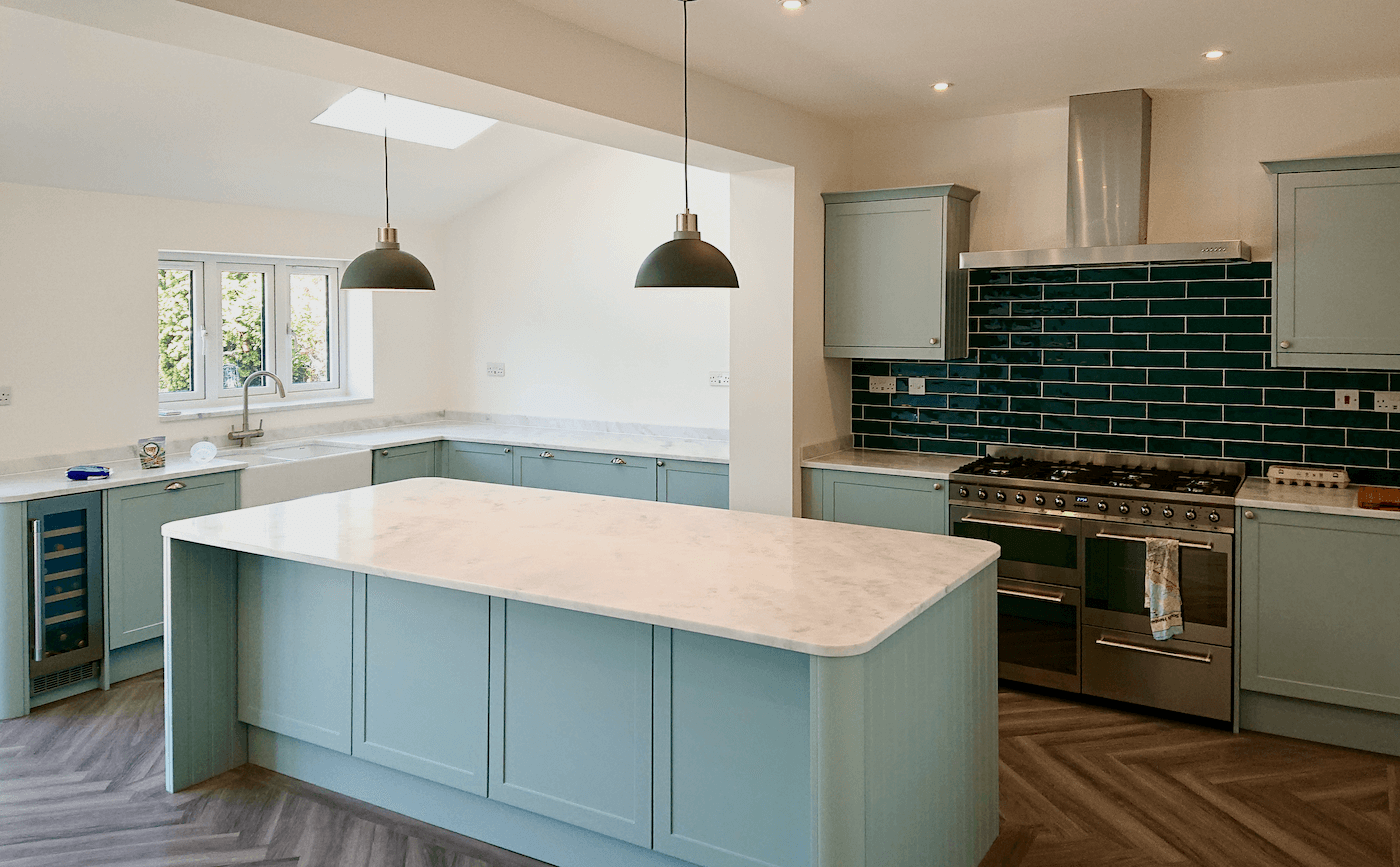 Modern kitchen in Westbury-on-Trym, Bristol with light blue cabinets, a marble island, black pendant lights, a stainless steel stove with a dark green tile backsplash, and a window with greenery outside.