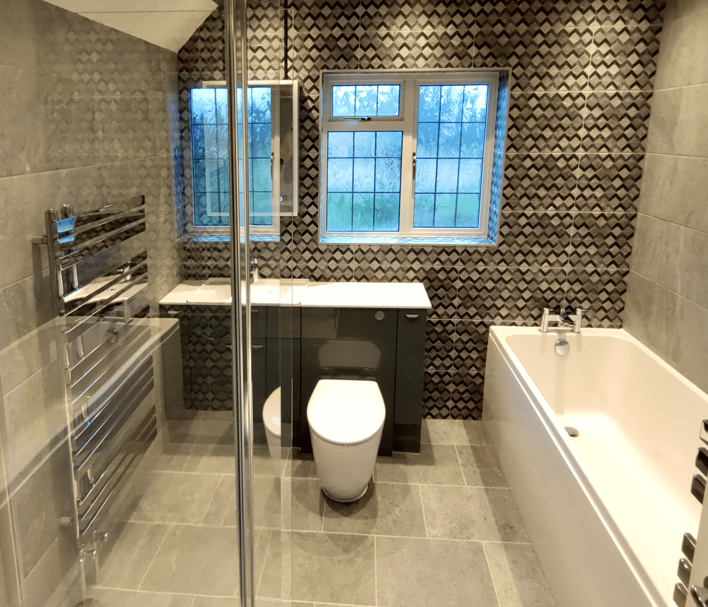 Modern bathroom in Stoke Bishop, Bristol with grey tiled floors and a textured, geometric patterned accent wall behind a window. The bathroom contains a white bathtub on the right, a black vanity with a white sink under the window, a mirror.