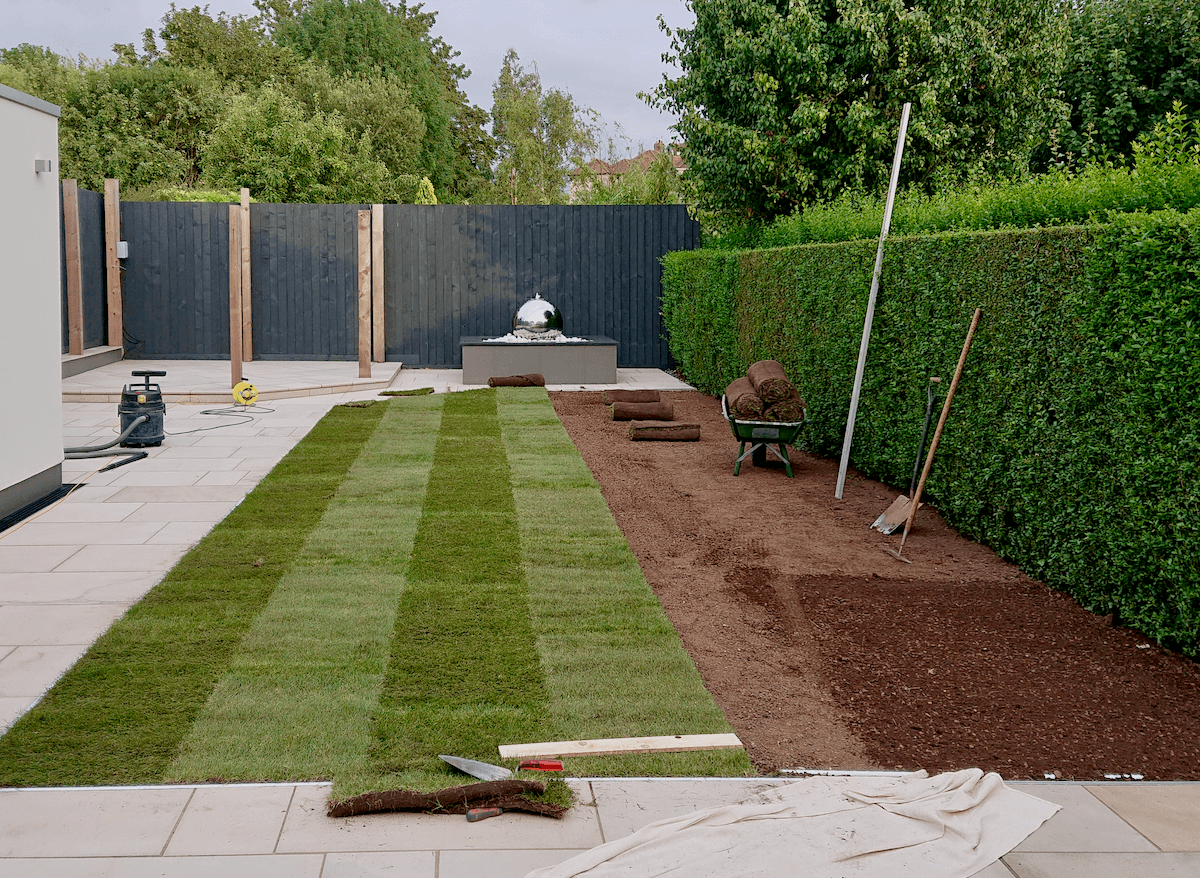 Contemporary garden in Filton Park, Bristol with freshly laid lawn, gardening tools, wheelbarrow with rolled sod, and a partially completed landscape project.