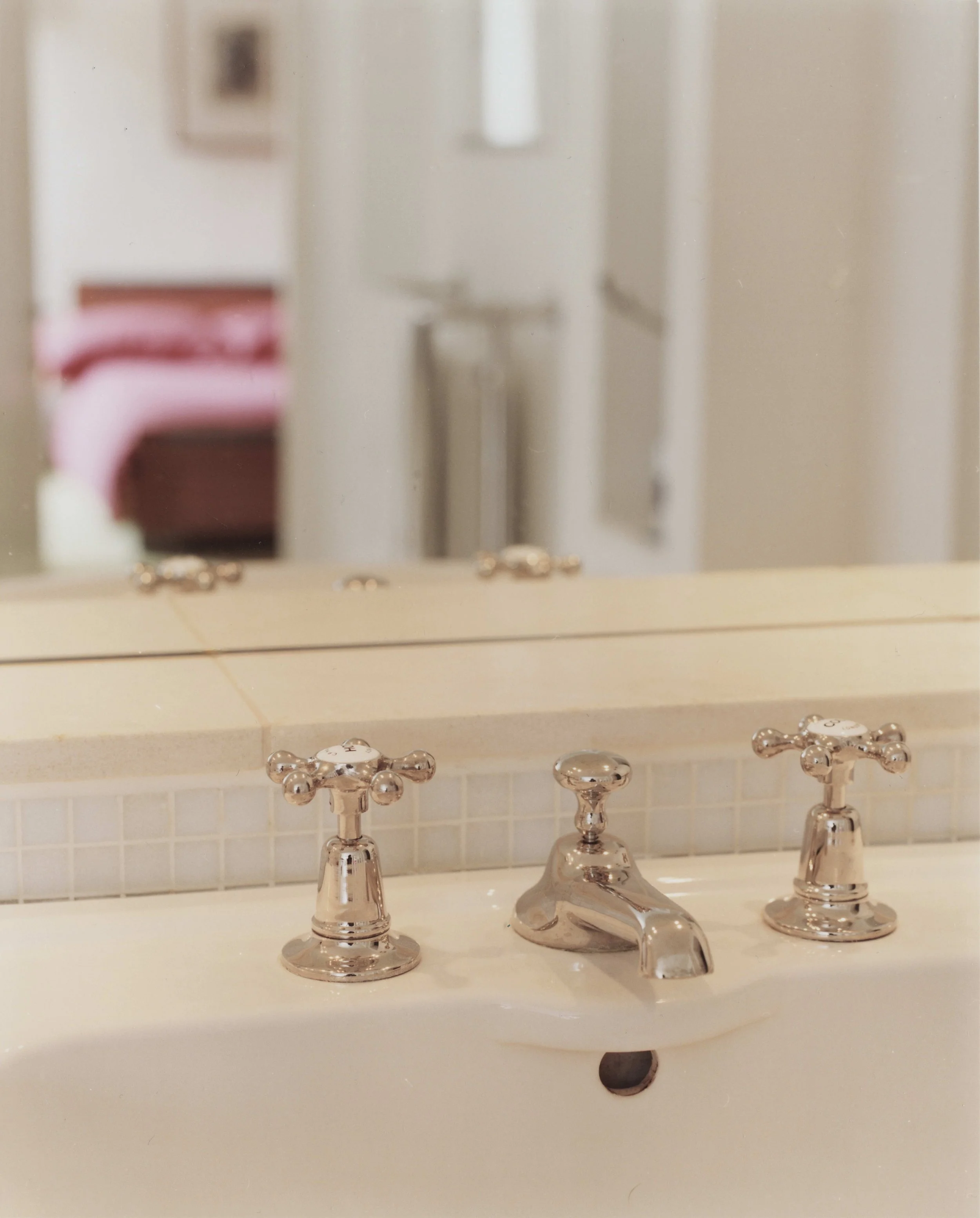 Close-up of a bathroom sink in a period building in Clifton, Bristol, with two taps, against a mirror and tiled backsplash, with a blurred bedroom visible in the background.
