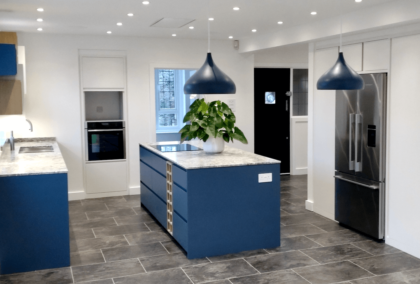 Modern luxury kitchen in Frenchay, Bristol with white cabinets, blue island, stainless steel refrigerator, black oven, and two large blue pendant lights over the island. A potted plant sits on the island.