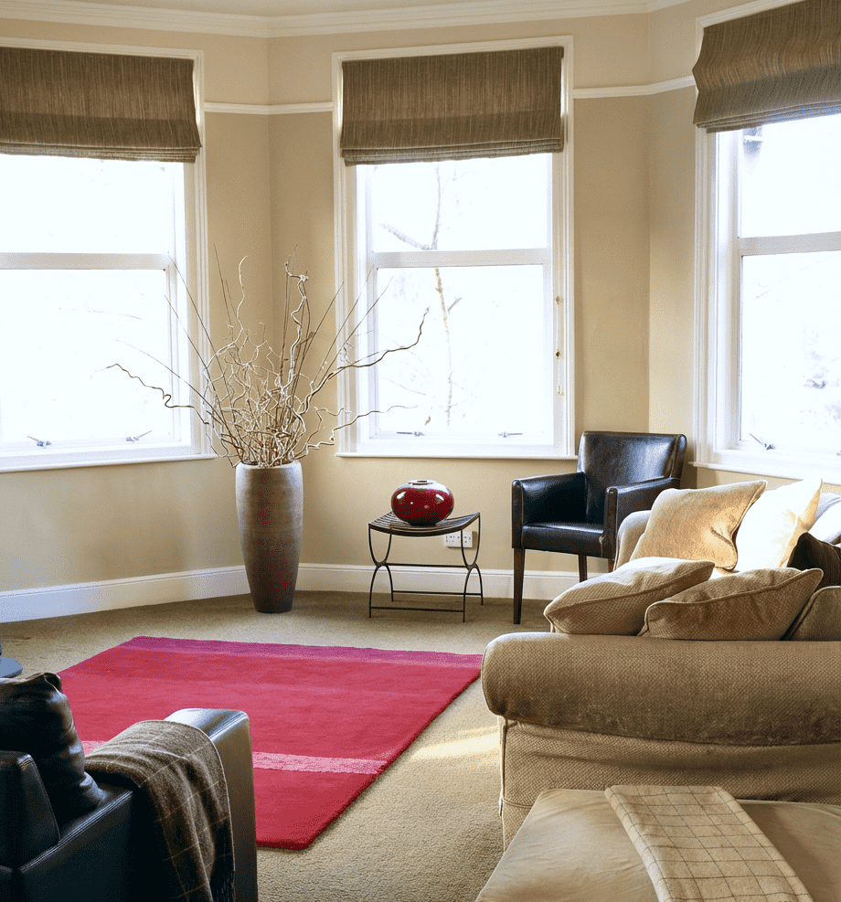 Living room in Cotham, Bristolwith three large windows, beige walls, a beige carpet, a red area rug, a tall vase with decorative branches, a black armchair, a beige armchair with pillows, a small table with a red decorative sphere, and window blinds.