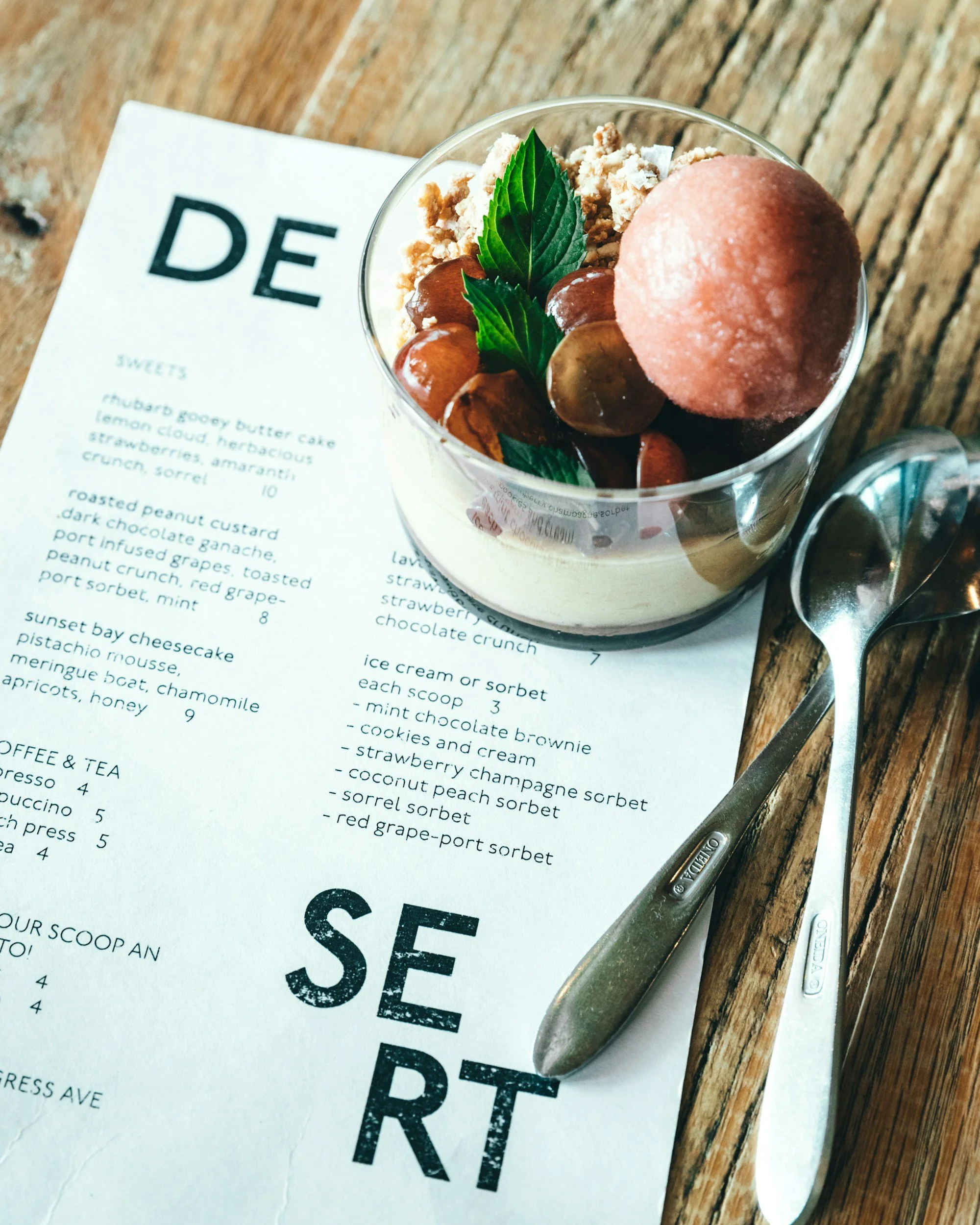A dessert glass with a pink sorbet, chocolate ganache, chocolate-covered almonds, and mint leaves on top, placed on a wooden table next to a spoon and dessert menu.