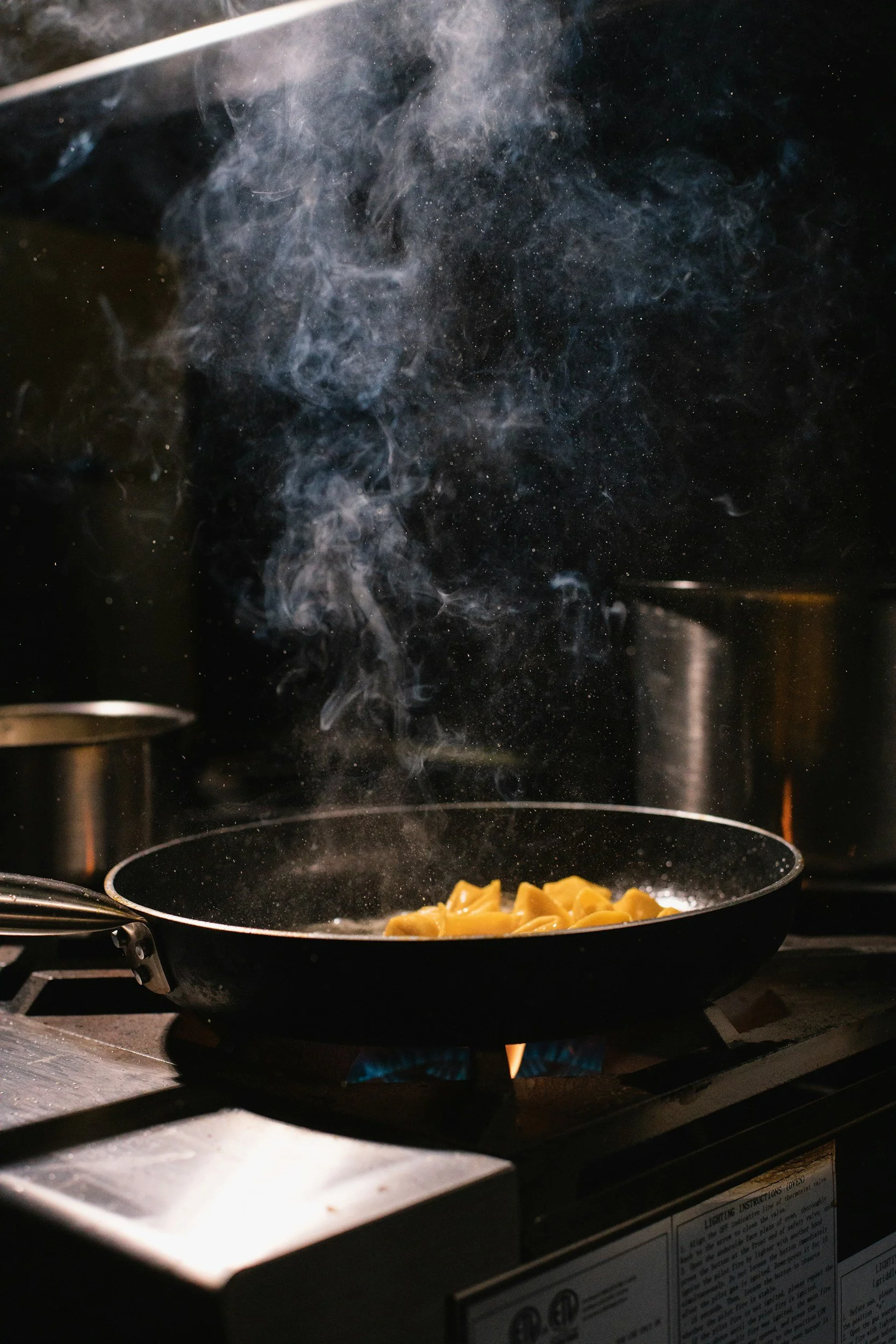 Frying pasta in a black skillet on a stove with blue flames visible underneath, with steam and smoke rising from the pan.