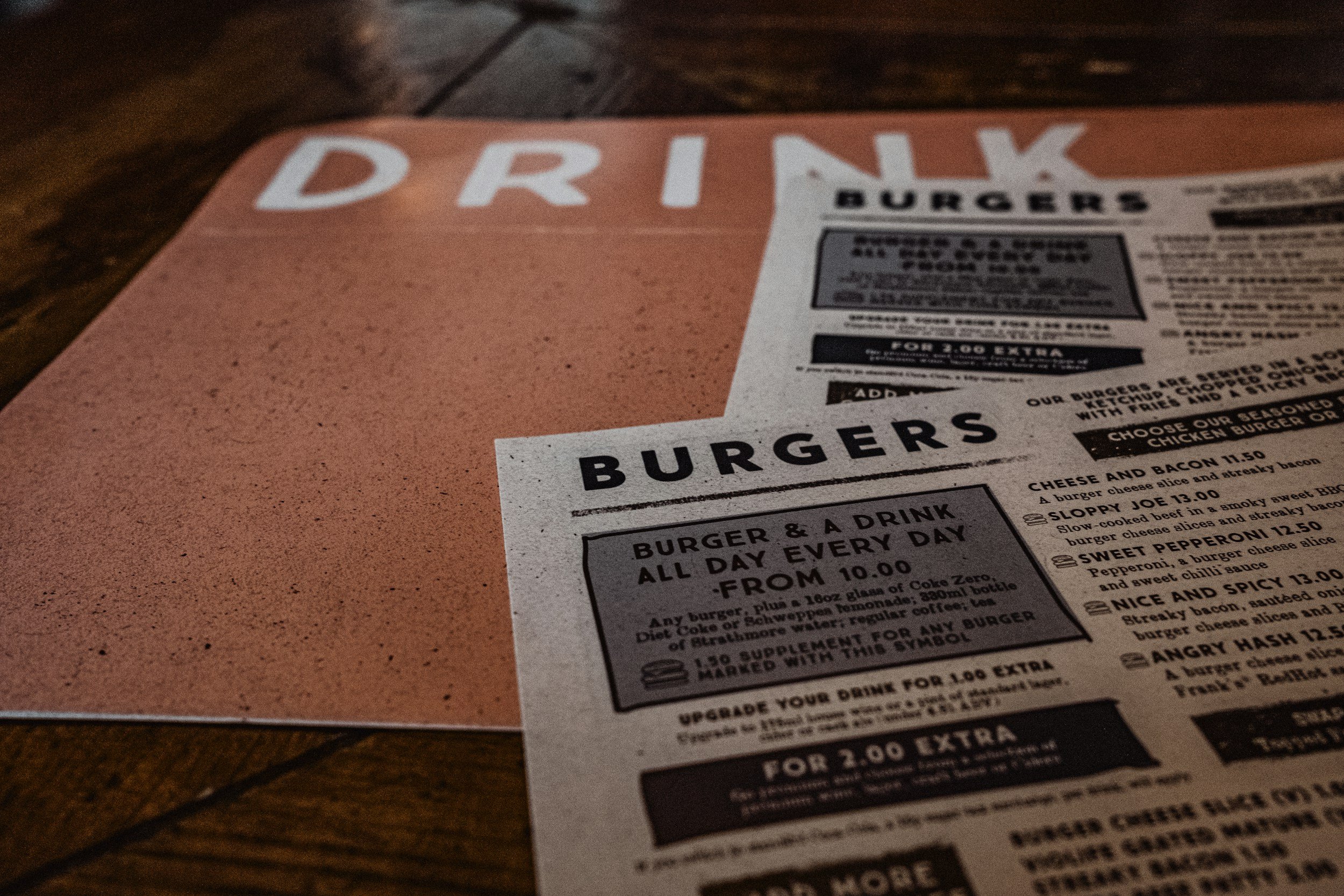 A restaurant menu on a wooden table with the word 'DRINK' partially visible at the top, and a section of burger options below.