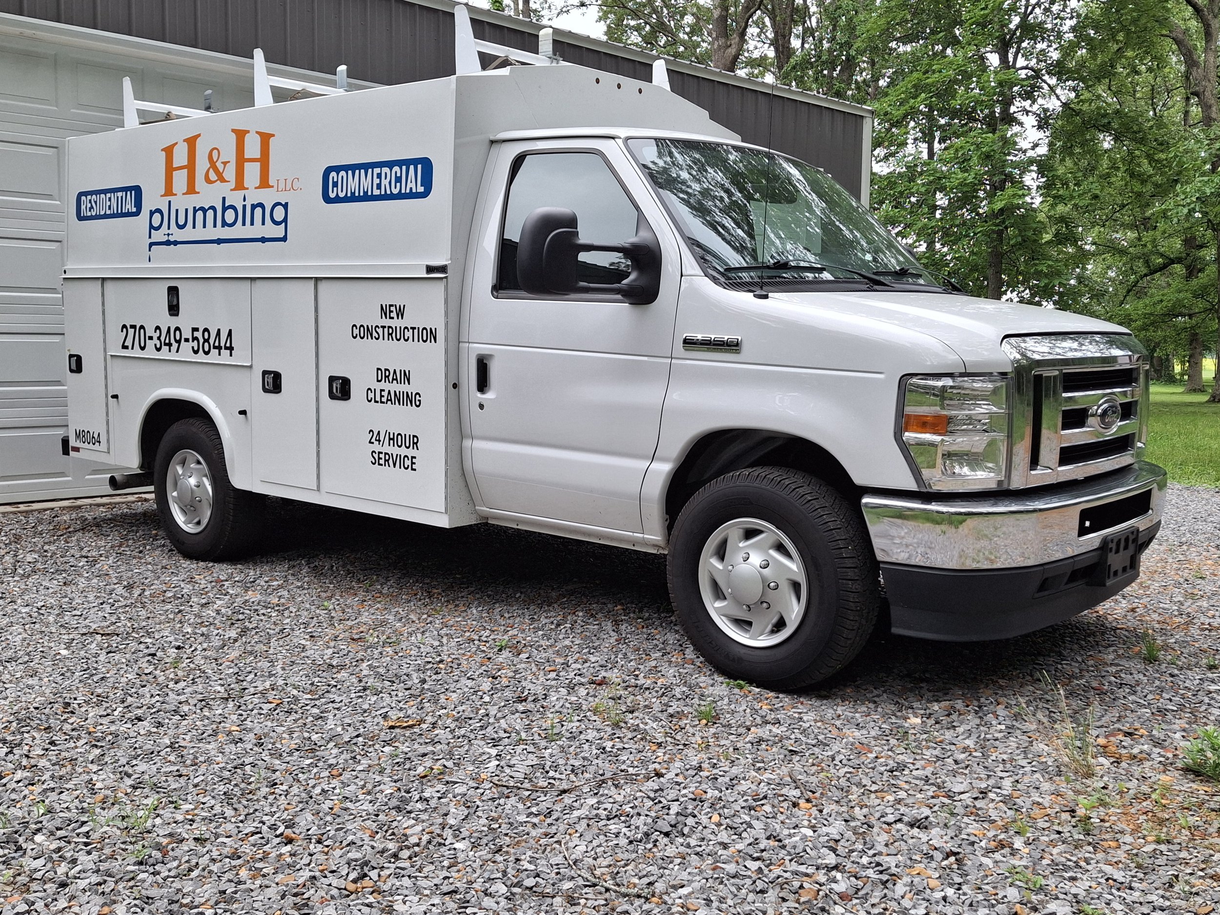 White service truck parked on gravel with green trees and a building in the background. The truck has signage for H&H Plumbing LLC, offering residential, commercial, and new construction plumbing services, including drain cleaning and 24-hour service.