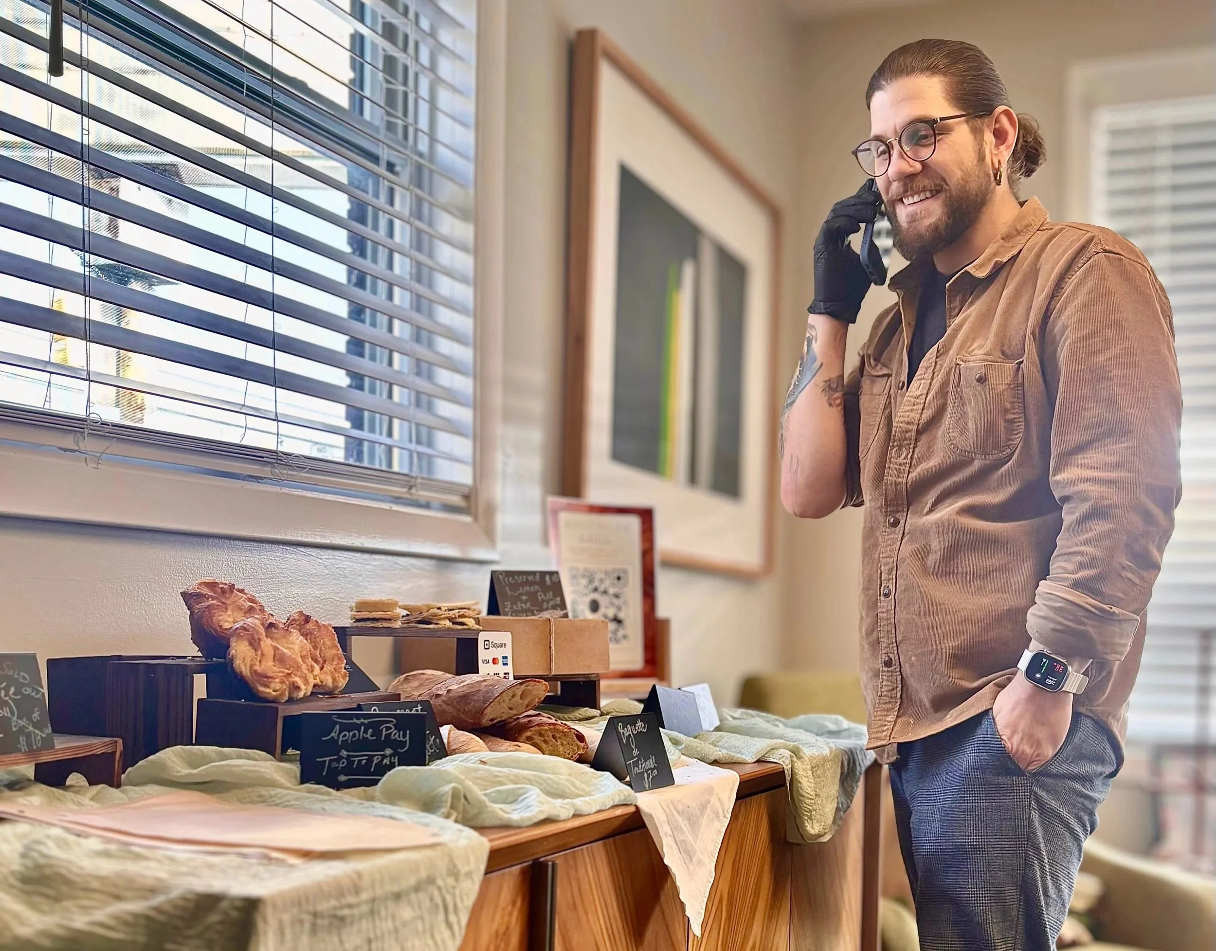 A man with glasses and a man bun is smiling and talking on the phone while browsing baked goods at a market stall. The stall has various bread and pastries on display with small signs and a QR code for payment. The setting is indoors near a window with blinds.