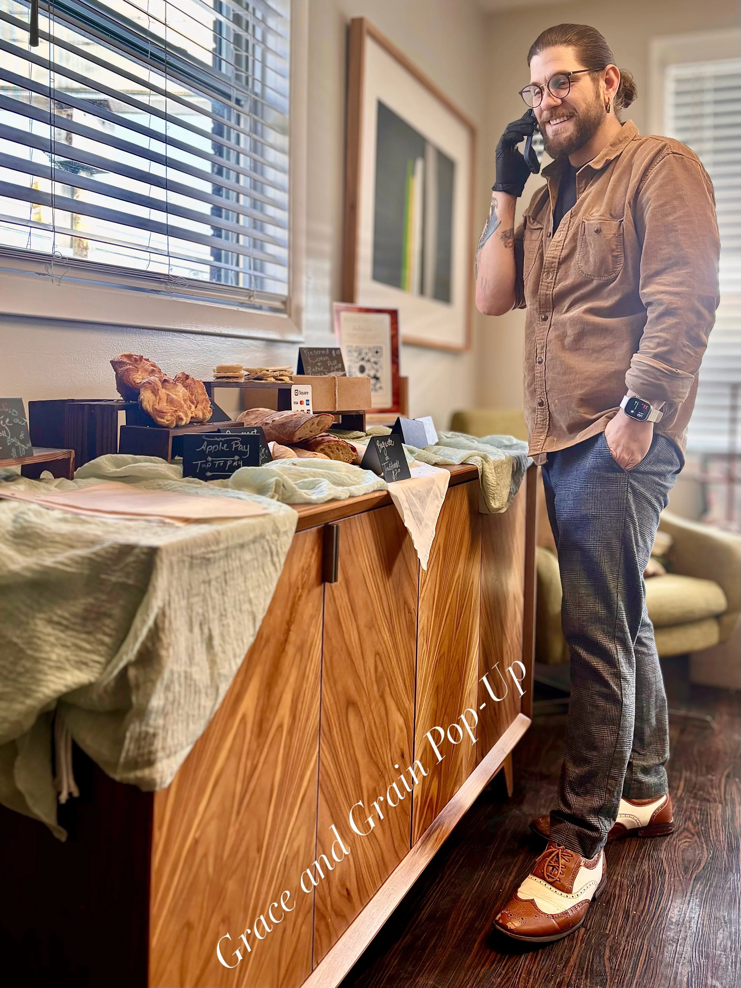 A man with glasses, tattoos, and a beard, wearing a brown shirt, plaid pants, and brown shoes, is talking on a cellphone at a small food stand or market display inside a cozy room with wooden floors and a window with blinds.