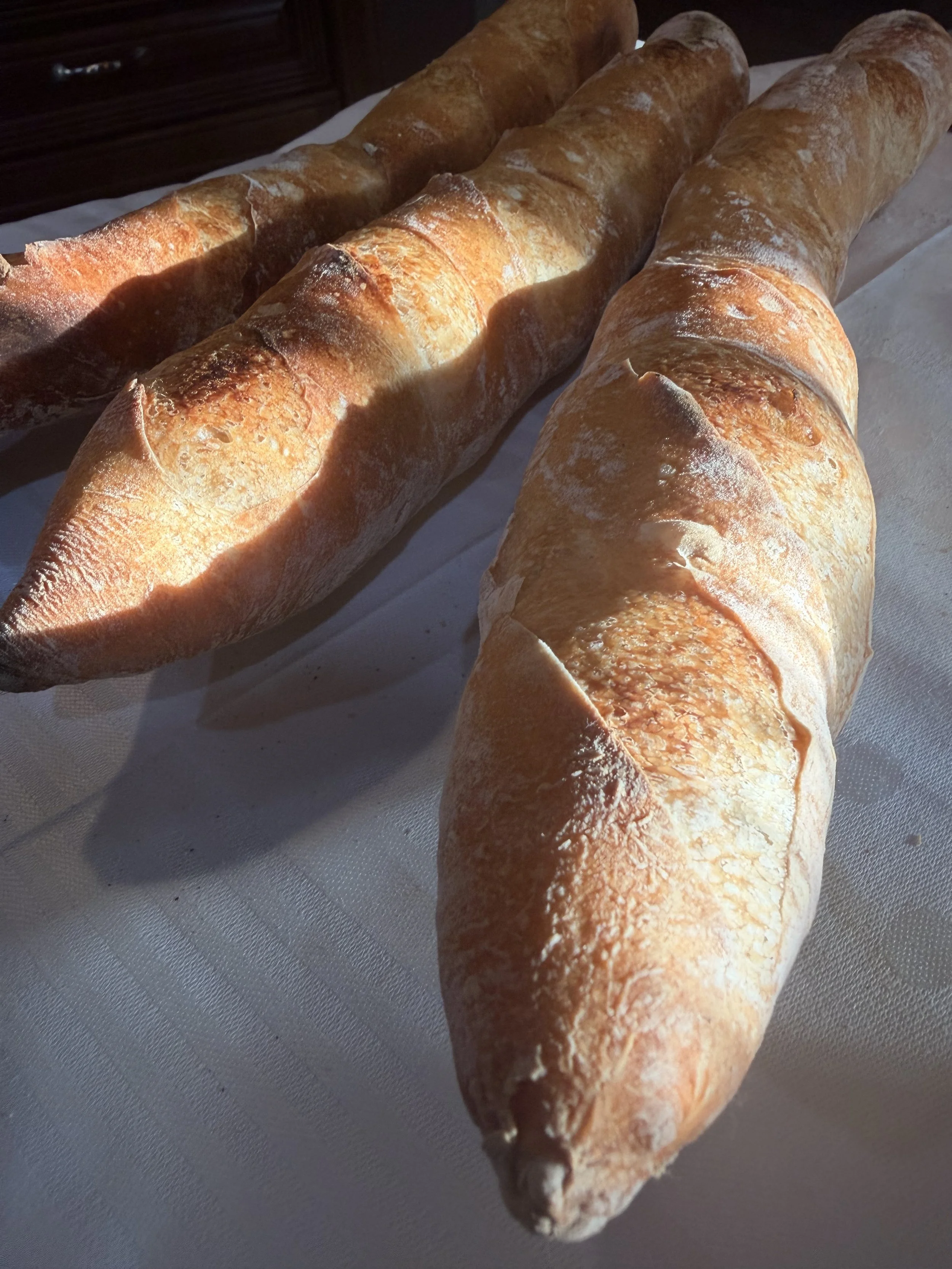 Three freshly baked baguettes with crispy crusts resting on a white surface, sunlight casting shadows.