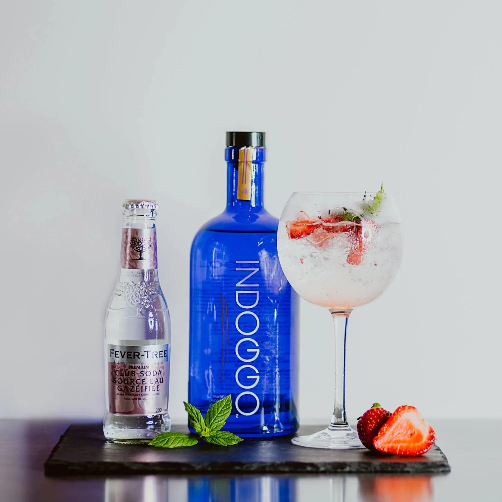 A blue bottle labeled 'Indoggo' rum, a small bottle of Fever-Tree tonic water, a glass of sparkling cocktail with strawberries and a lime wedge, on a black serving board with strawberries and mint leaves, against a plain white background.