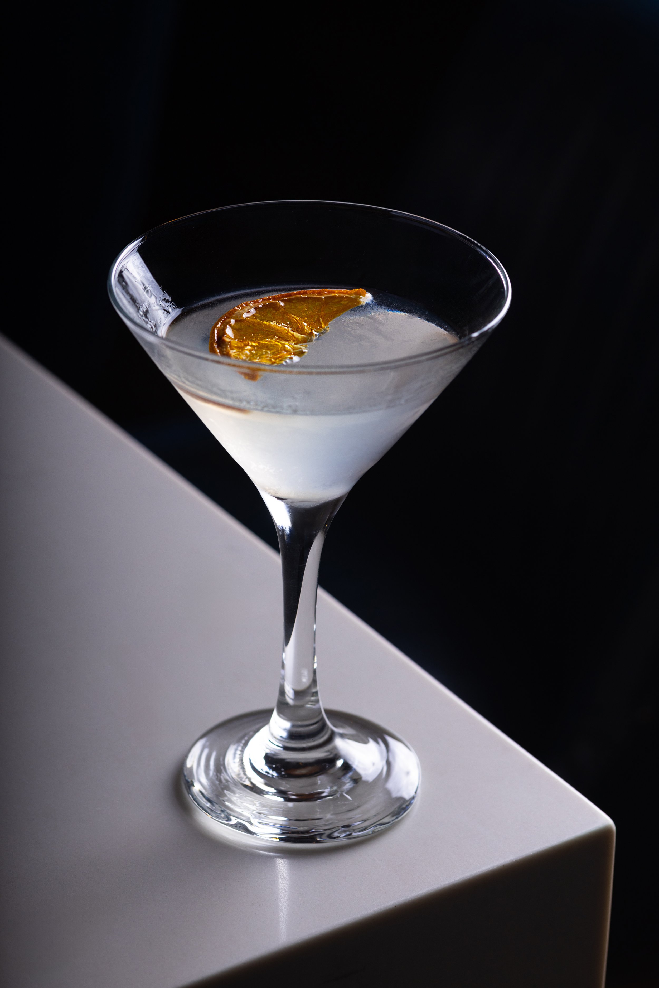 Southside Shake's olive oil washed martini served in a martini glass and garnished with a dried orange slice, placed on a white surface of the bar against a dark background.