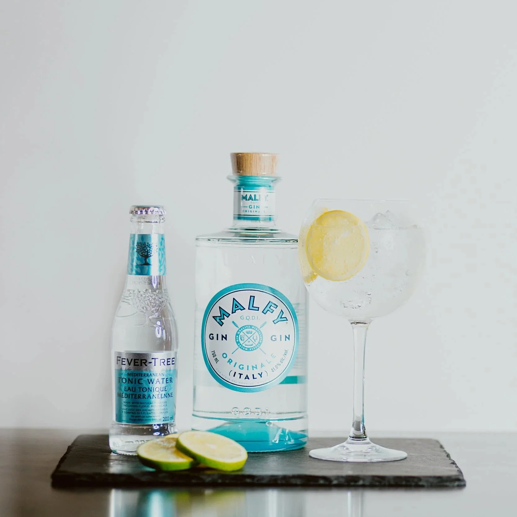 Bottle of Malfy gin, Fever-Tree tonic water, and a gin and tonic drink with lemon slices on a wooden surface against a plain white background.