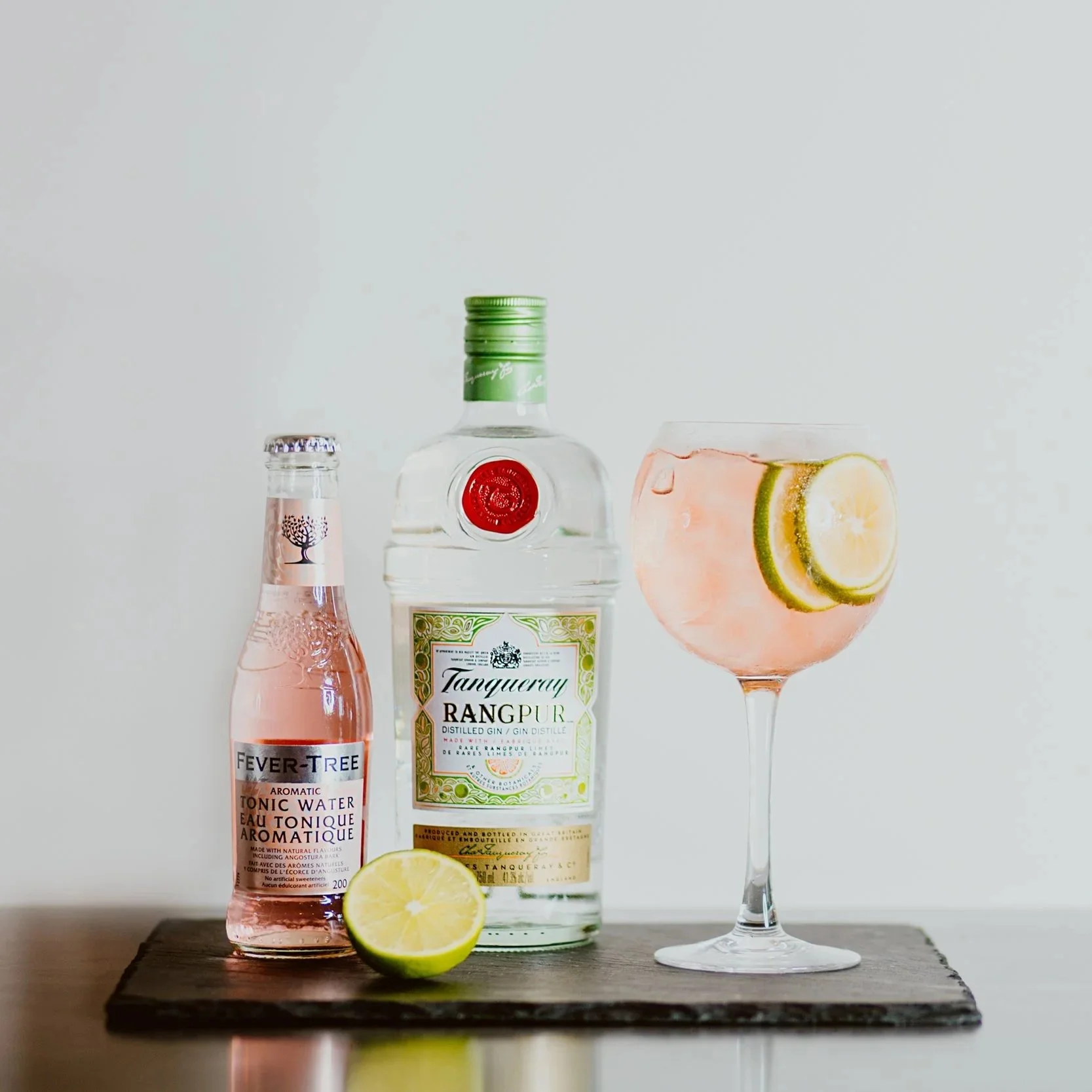 A bottle of Tanqueray Rangpur gin, a bottle of Fever-Tree pink tonic water, and a cocktail glass with a pink drink garnished with lemon and lime slices, all placed on a dark wooden surface with a lemon half in front.