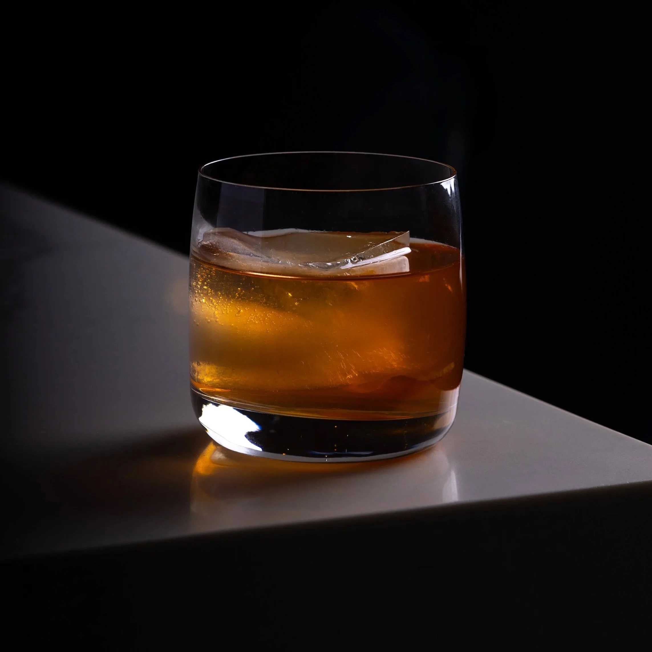 A glass of whiskey with a large ice cube, Southside Shake's Smoke & Honey Old Fashioned, on the white bar top against a black background.