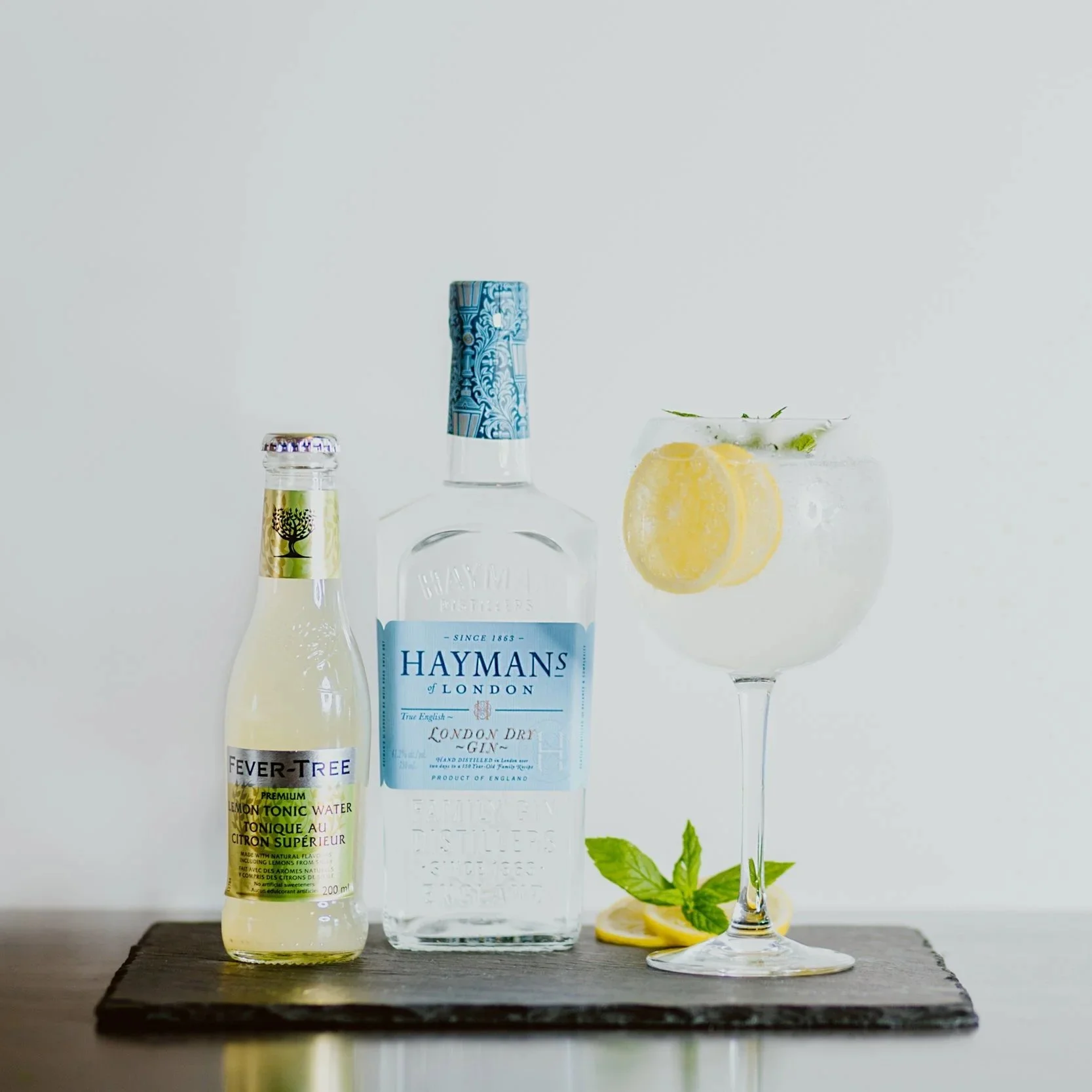 A bottle of Haymans London Dry Gin, a bottle of Fever-Tree Lemon Tonic Water, and a gin and tonic in a glass with lemon slices and mint leaves on a dark wooden surface against a plain white background.