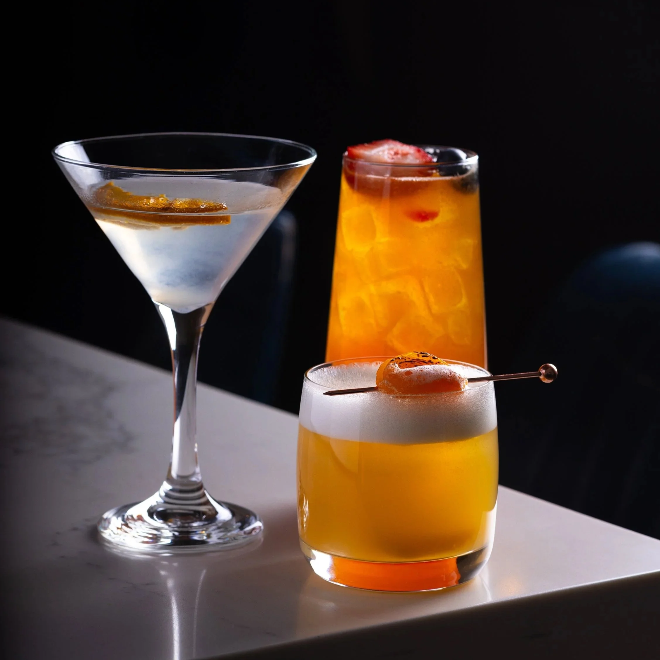 Three cocktails on a white surface against a black background. The left cocktail is in a martini glass with clear liquid and garnish. The center cocktail is in a tall glass with orange liquid and ice, garnished with a strawberry. The right cocktail is in a short glass with yellowish liquid, foam on top, and a skewer with a fruit piece.