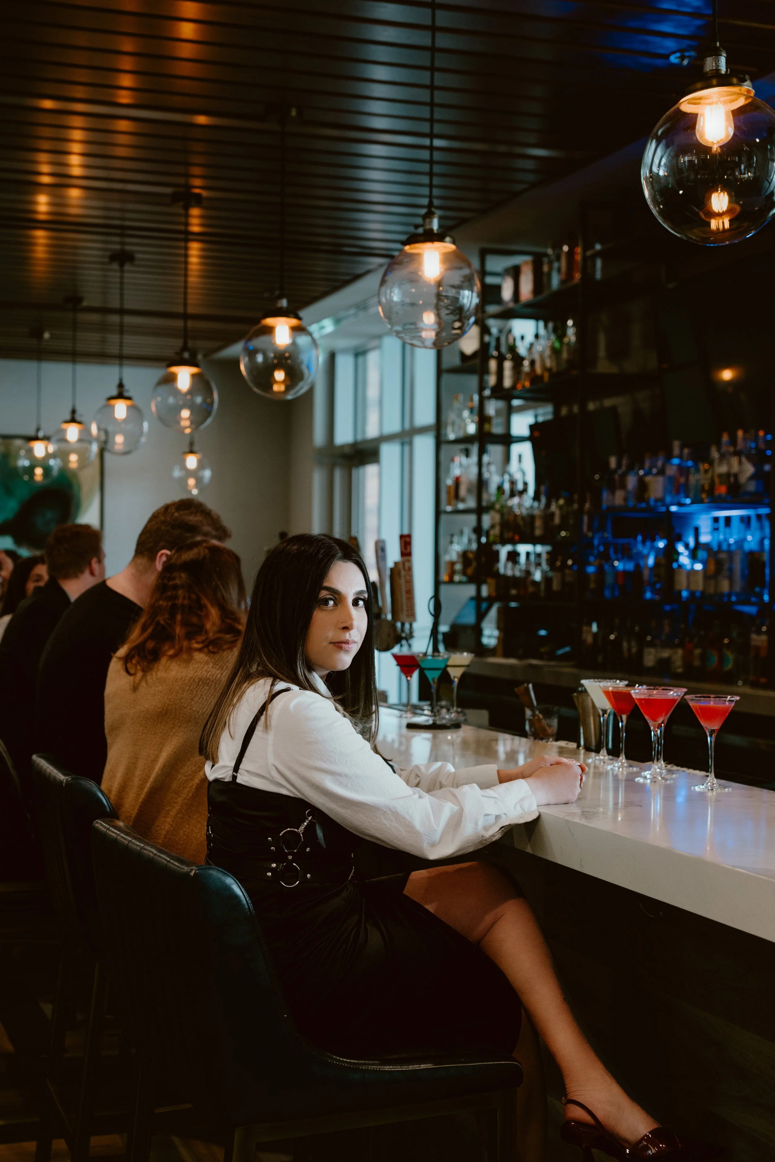 A woman sitting at Southside Shake bar with several colourful cocktails in front of her, in the dimly lit bar with hanging globe lights and shelves filled with gin bottles in the background.