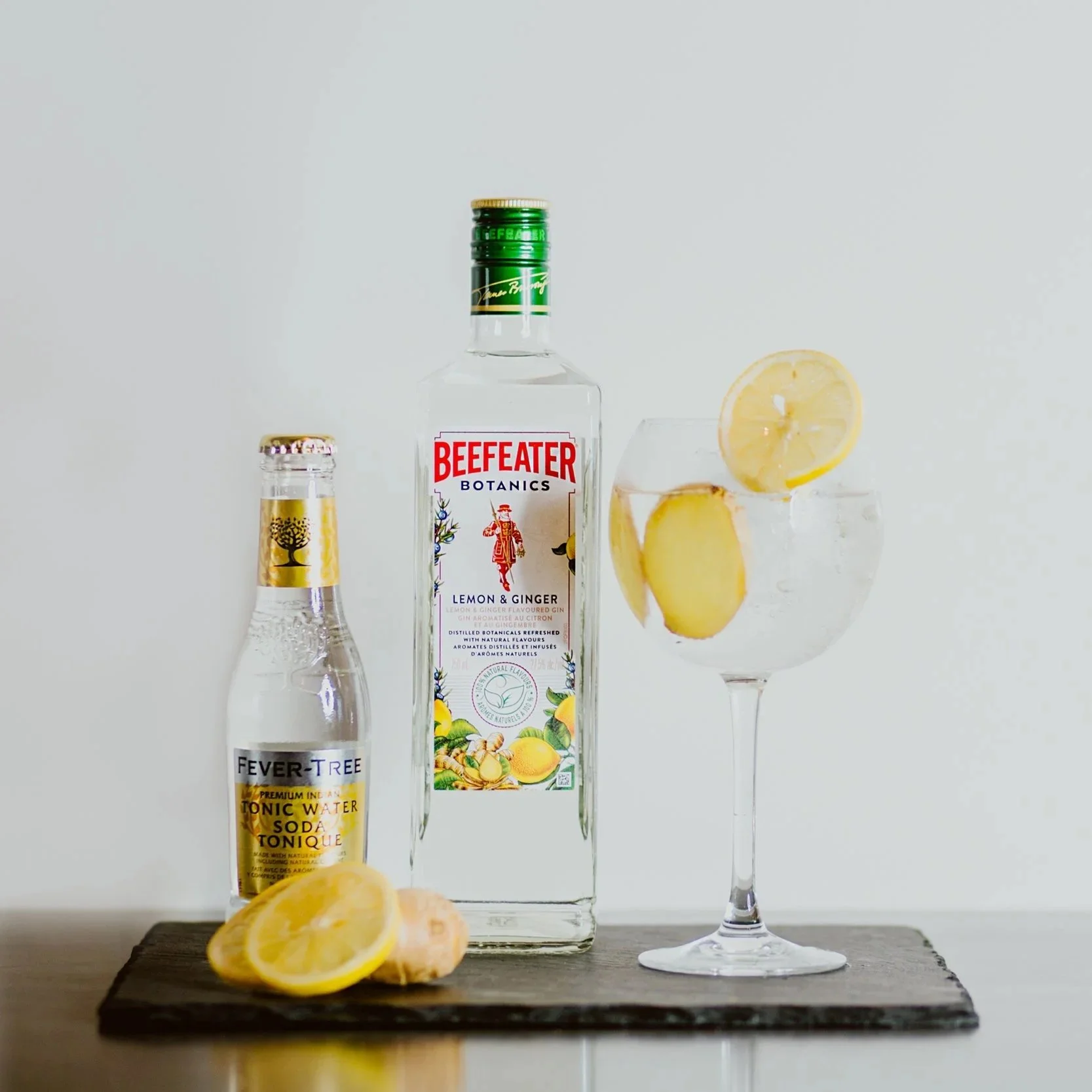 A bottle of Beefeater Botanics Lemon & Ginger gin, a Fever-Tree tonic water bottle, and a glass of gin and tonic garnished with lemon slices are arranged on a wooden surface against a plain background.