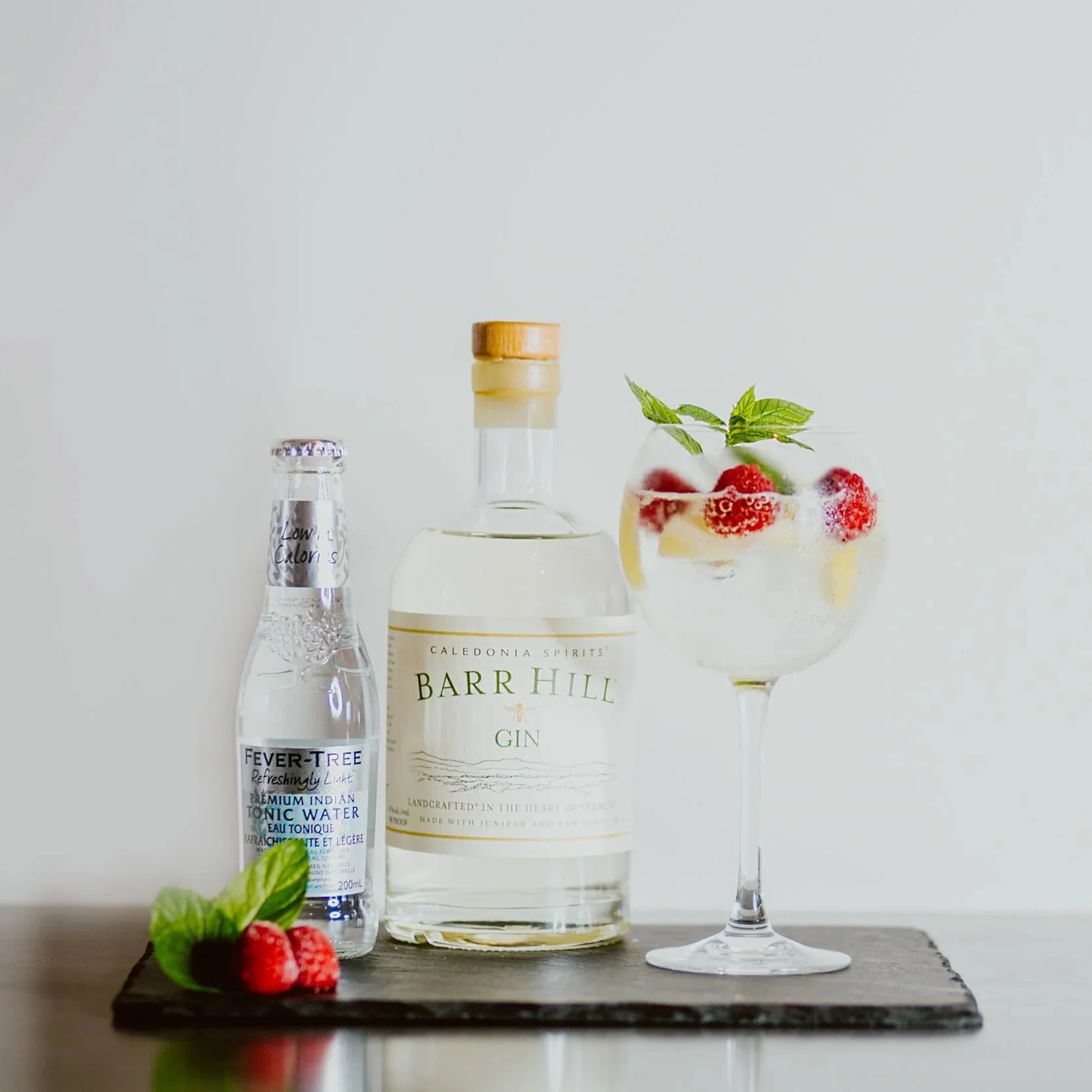 A bottle of Barr Hill gin, a bottle of Fever-Tree tonic water, and a large glass of gin and tonic with strawberries, mint, and ice on a black tray.