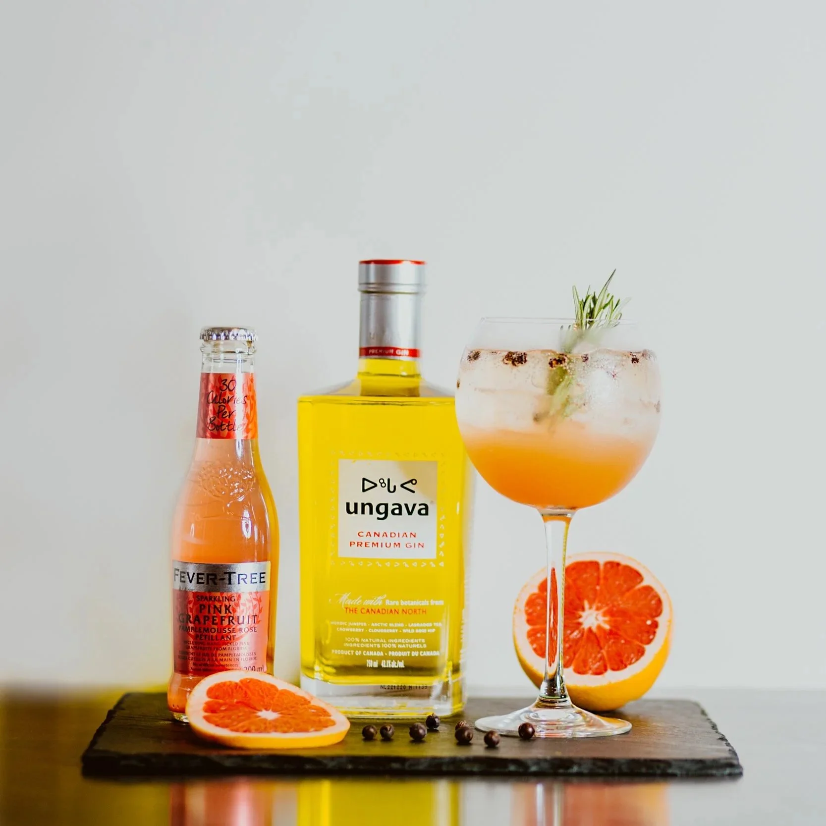 A bottle of Ungava Canadian gin, a bottle of Fever-Tree pink grapefruit soda, a grapefruit half, a cocktail glass with a pink drink garnished with herbs, on a wooden surface against a plain background.