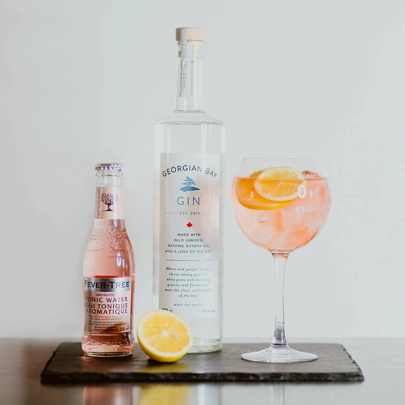 A glass of pink gin and tonic with lemon slices, a bottle of Georgian Bay gin, a bottle of Fever-Tree tonic water, and a lemon half on a dark wooden surface against a plain light background.