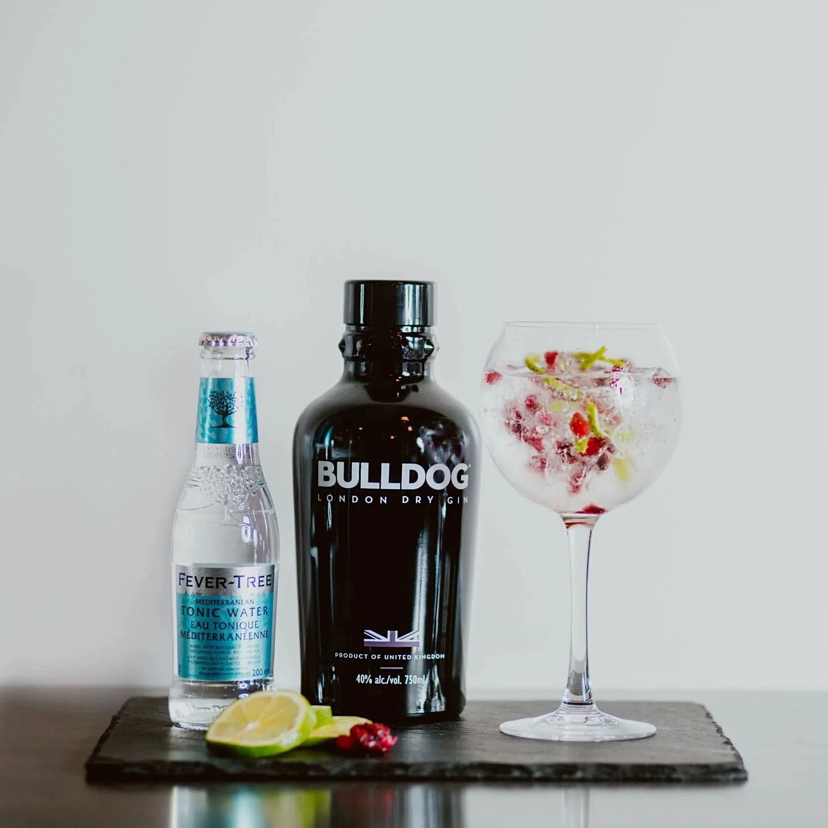 A bottle of Bulldog London Dry Gin, a bottle of Fever-Tree tonic water with lemon and pomegranate slices in a glass of gin and tonic on a wooden board.
