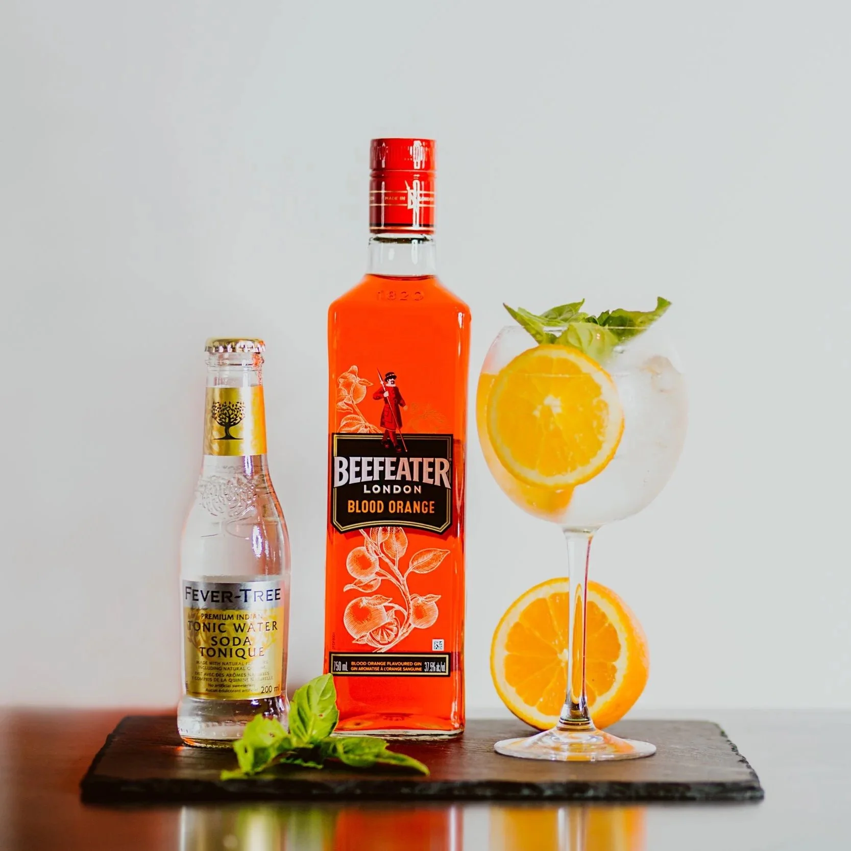 A bottle of Beefeater London Blood Orange gin, a small bottle of Fever-Tree tonic water, and a cocktail glass with ice, orange slices, and mint leaves on a wooden surface.