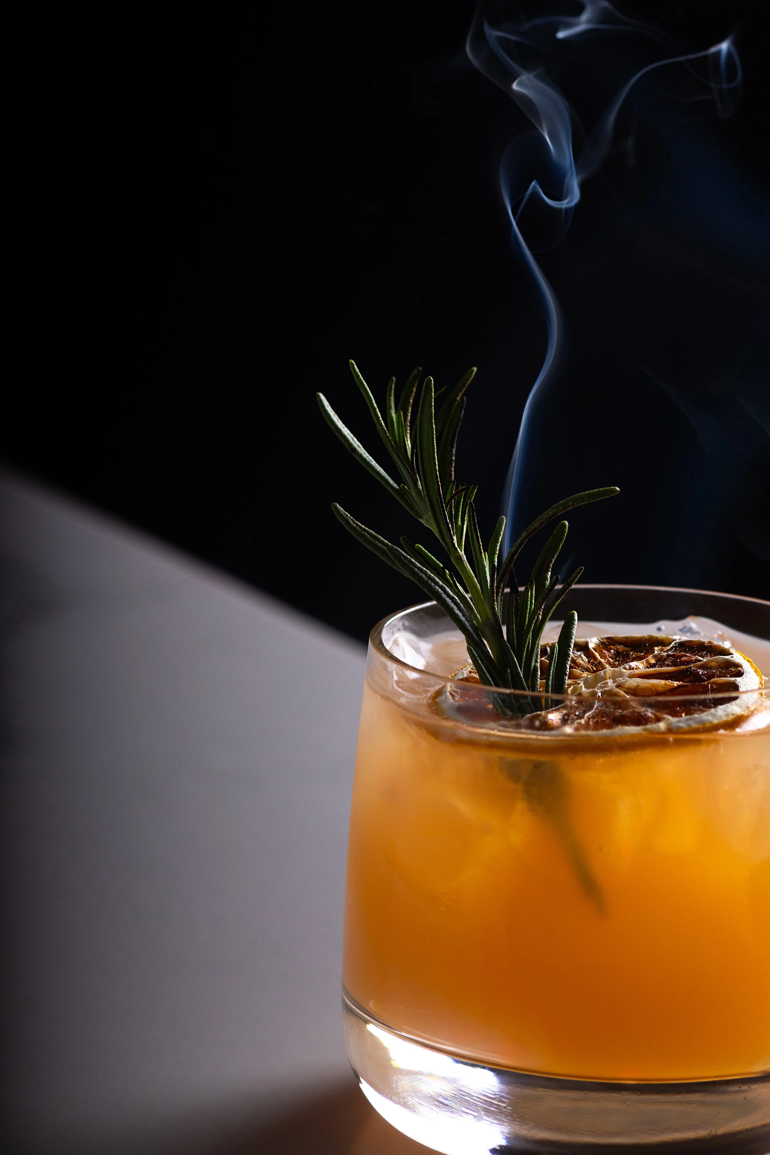 Southside Shake's Ginny Hop cocktail - An animated orange cocktail garnished with a sprig of rosemary and a dried citrus slice, with a lemon twist, on a dark background with a hint of white surface.