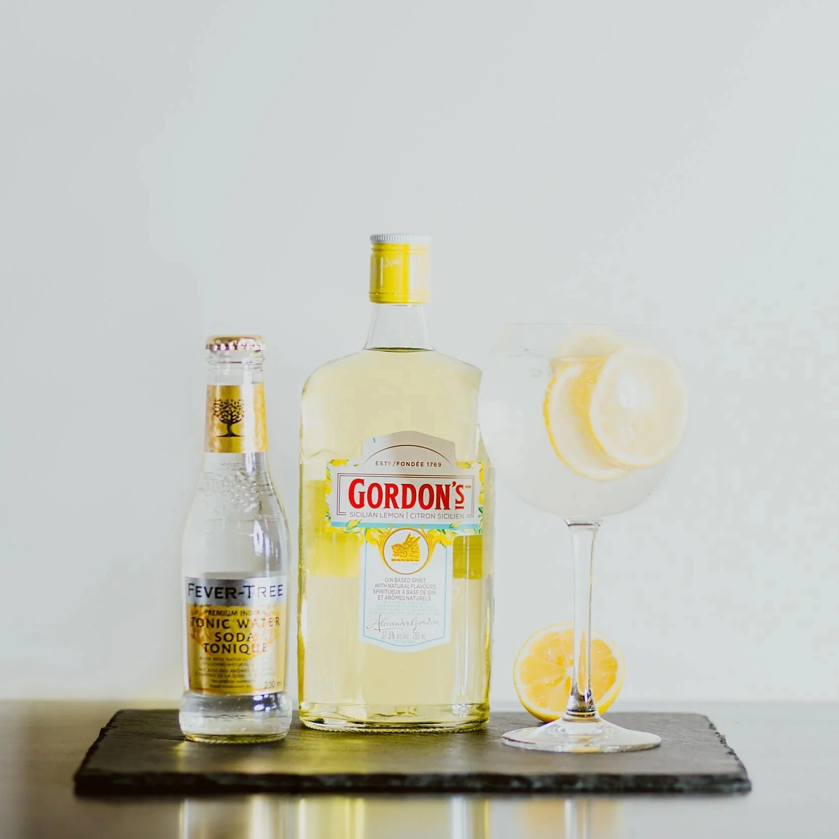 A bottle of Gordon's Sicilian Lemon gin, a bottle of Fever-Tree tonic water, and a cocktail glass with lemon slices on a wooden surface.