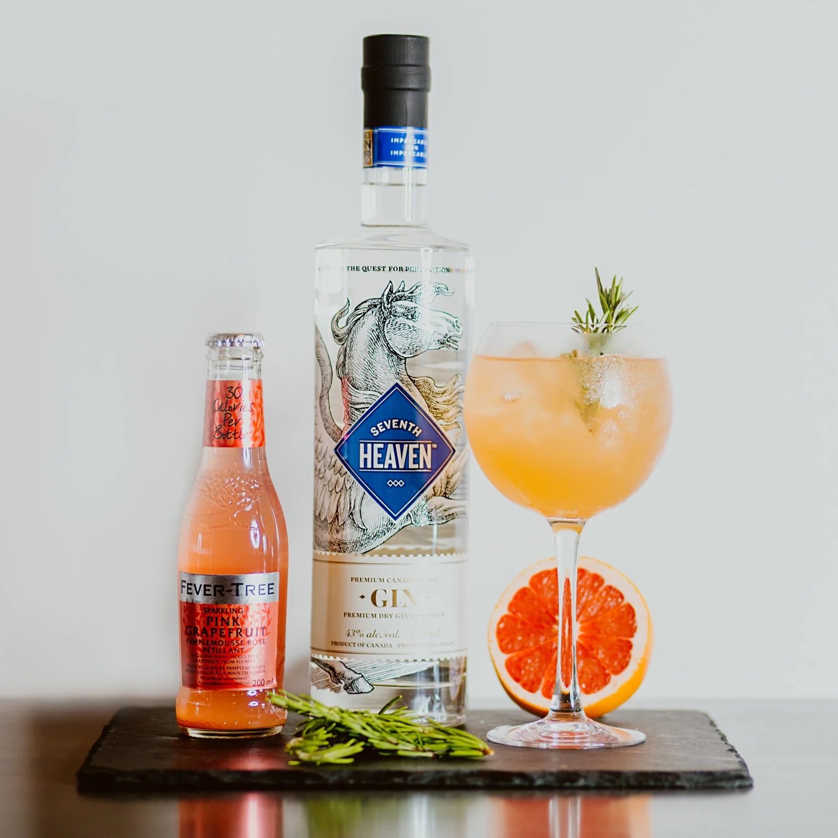 A cocktail in a stemmed glass garnished with a sprig of rosemary, with a half grapefruit and a bottle of Seventh Heaven gin and a bottle of Fever-Tree pink grapefruit tonic water on a wooden surface.