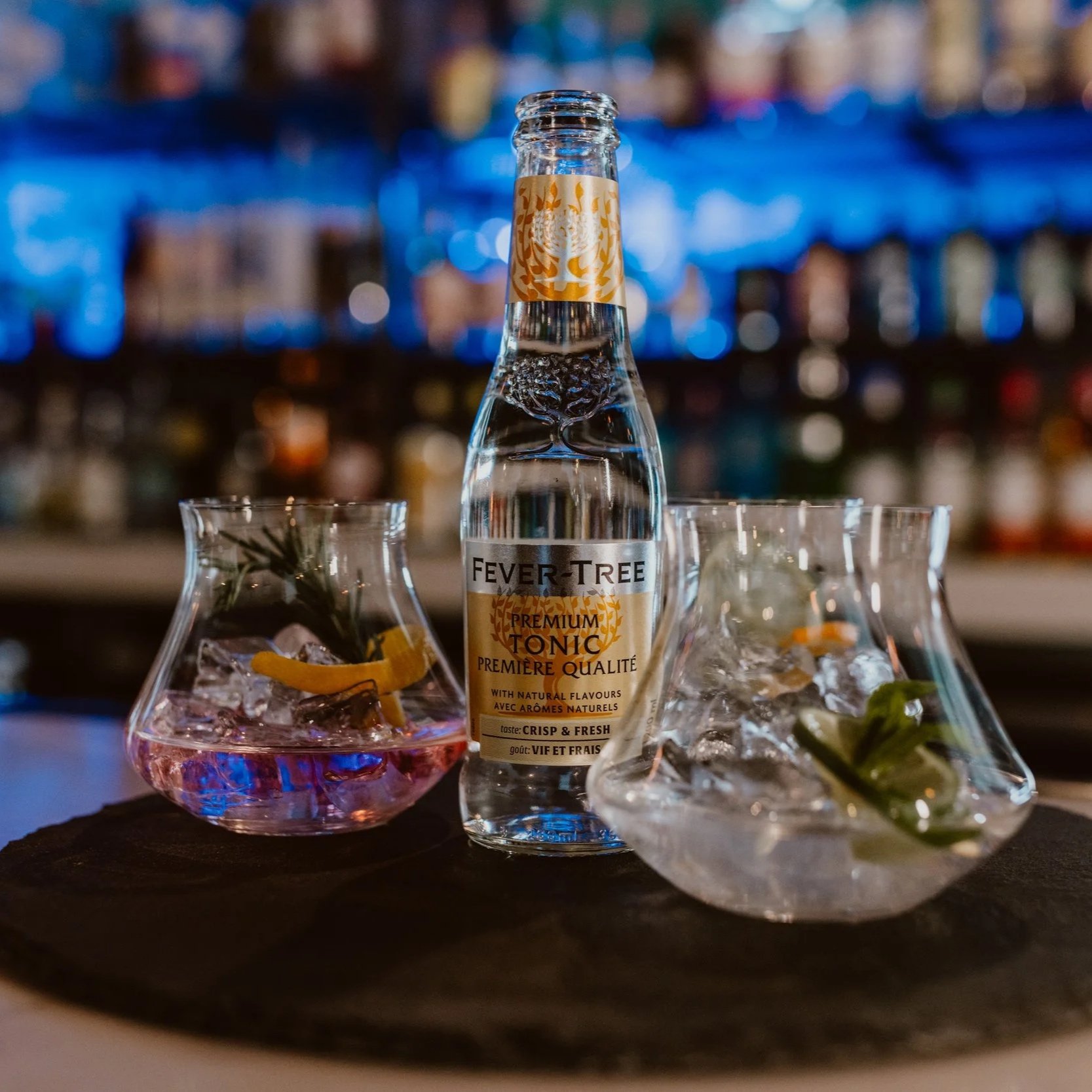 Southside Shake's Gin Flight served with a bottle of Fever-Tree premium tonic water flanked by three glasses with ice, lemon, and herbs on a bar top.