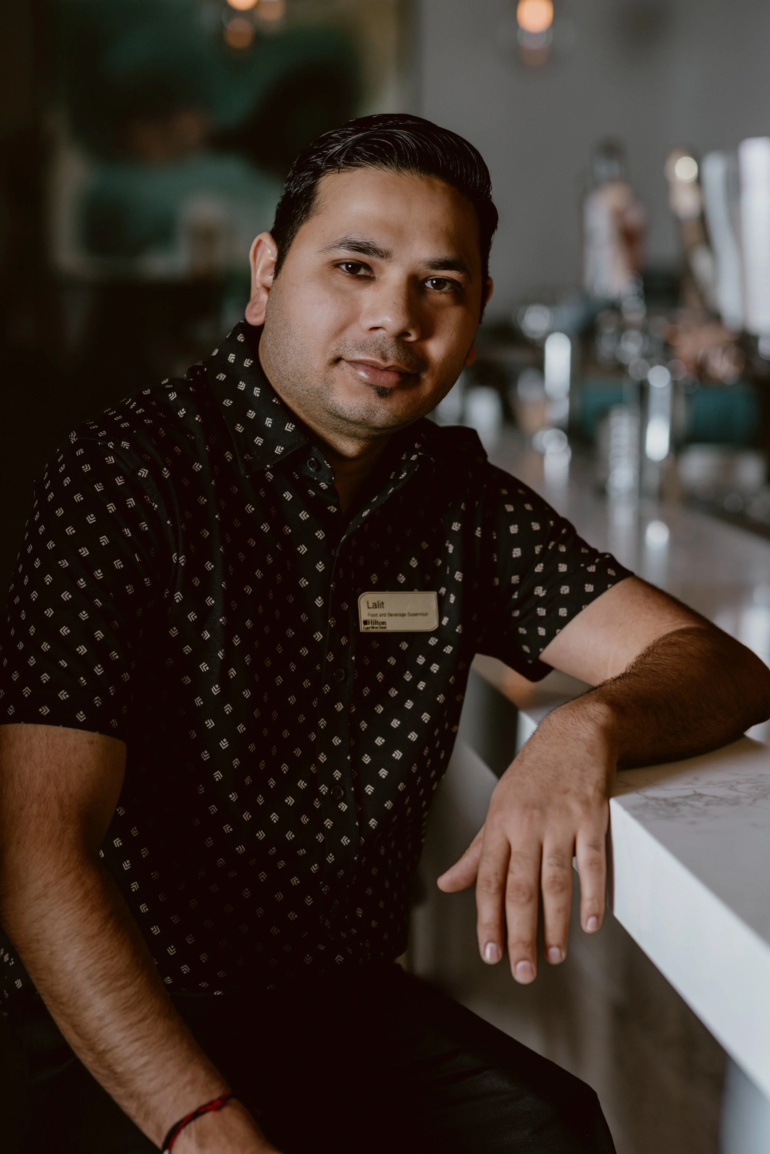 Head bartender, Lalit,  with short black hair wearing a black patterned shirt with a name tag, sitting at a Southside Shake bar  with a white marble surface, in a dimly lit environment.