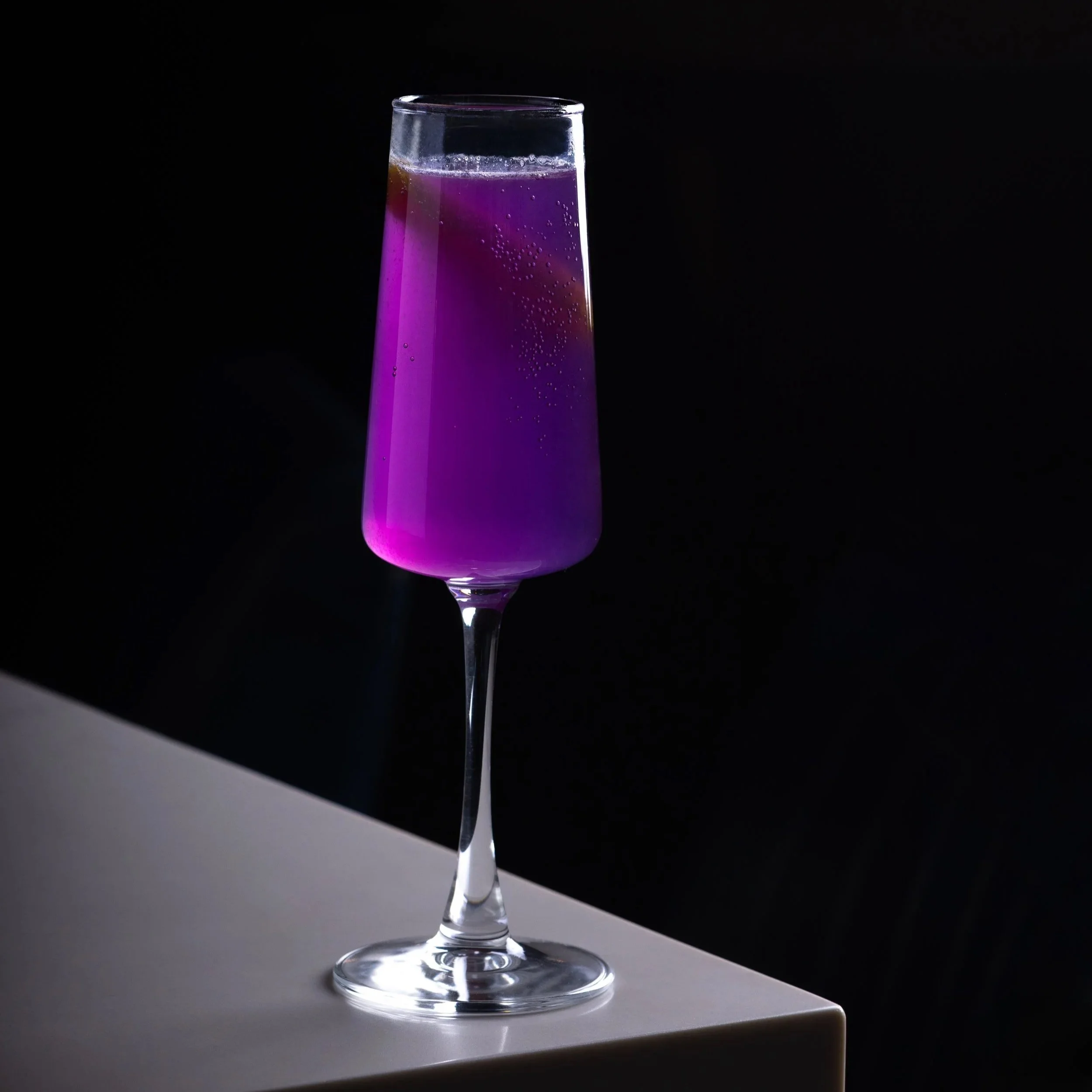 A tall champagne glass filled with a purple gin cocktail, Royal Flute, placed on the white bar top of Southside Shake with a black background.