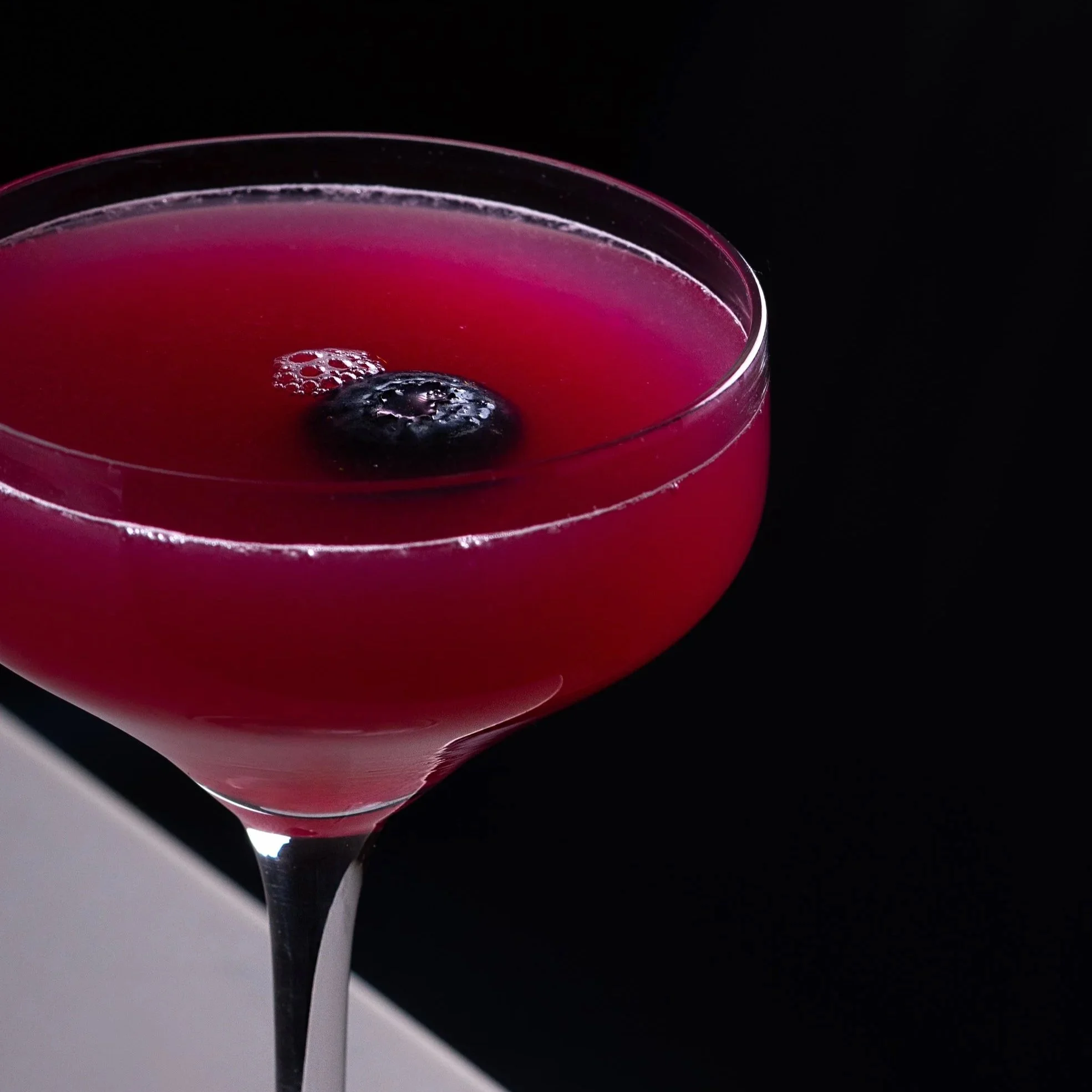 Southside Shake's Blueberry Delight gin cocktail - a pink cocktail with a blueberry garnish in a coupe glass against a black background.