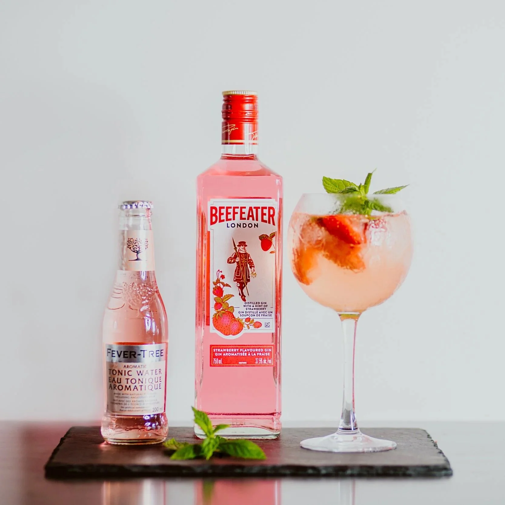 A bottle of Beefeater Gin with strawberry flavor, a small bottle of Fever-Tree strawberry tonic water, and a strawberry cocktail garnished with mint leaves on a black slate serving board.