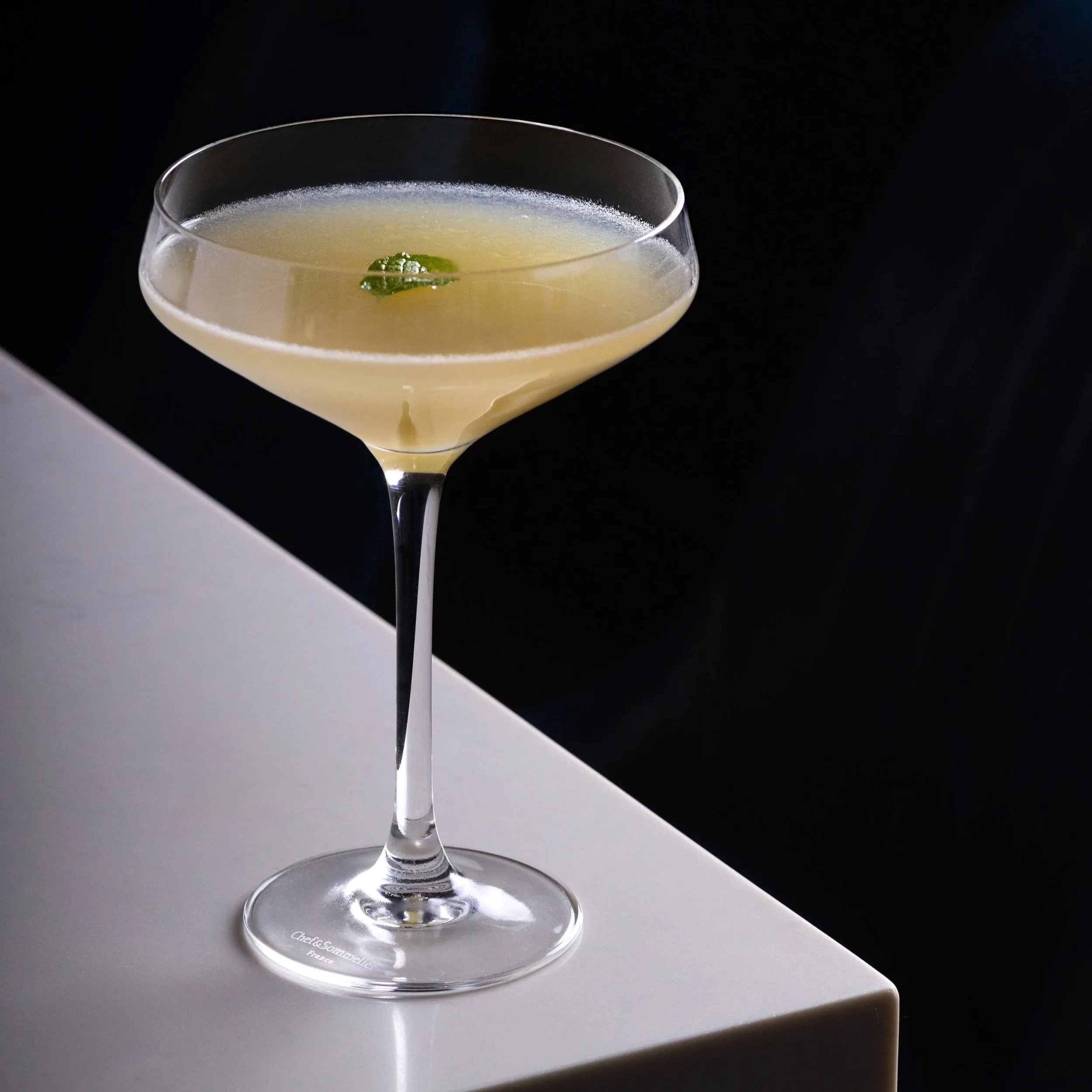 Southside Shake's namesake cocktail in a coupe glass with a green garnish on top, set on the white bar top with a dark background.