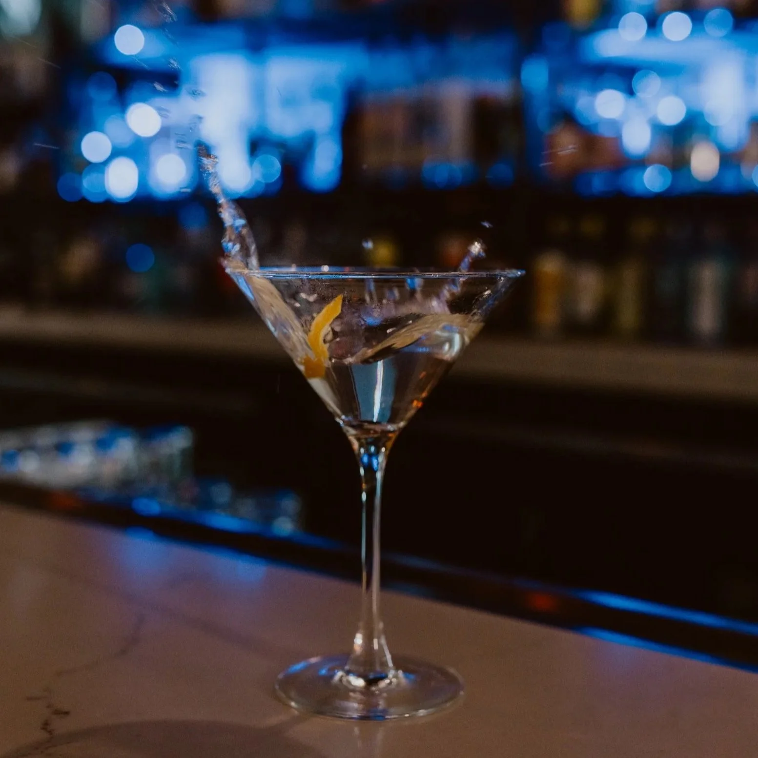 A martini glass with a lemon twist garnish, with liquid splashing, set against the blurred bar background of Southside Shake.