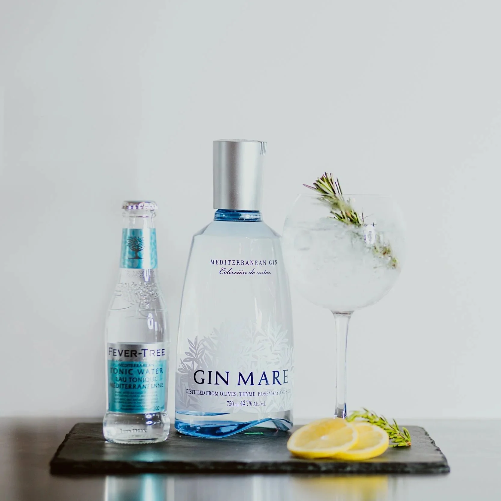 A bottle of Mediterranean gin, a bottle of Fever-Tree tonic water, and a large glass of gin and tonic garnished with rosemary, along with lemon slices and rosemary sprigs, all placed on a dark wooden surface against a plain white background.