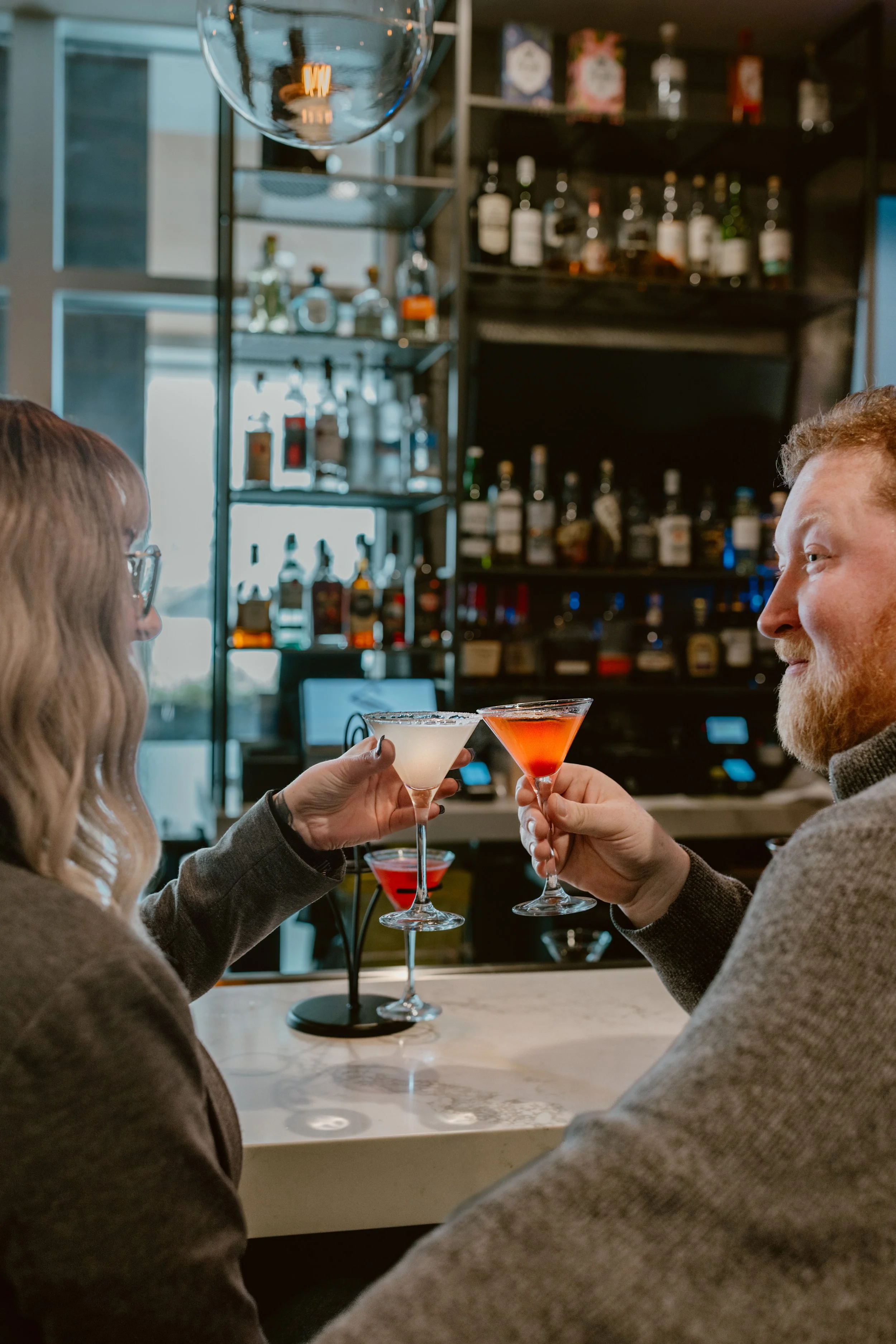 A man and woman clinking 2 tiny tini glasses at Southside Shake bar, with shelves of various gin bottles in the background.