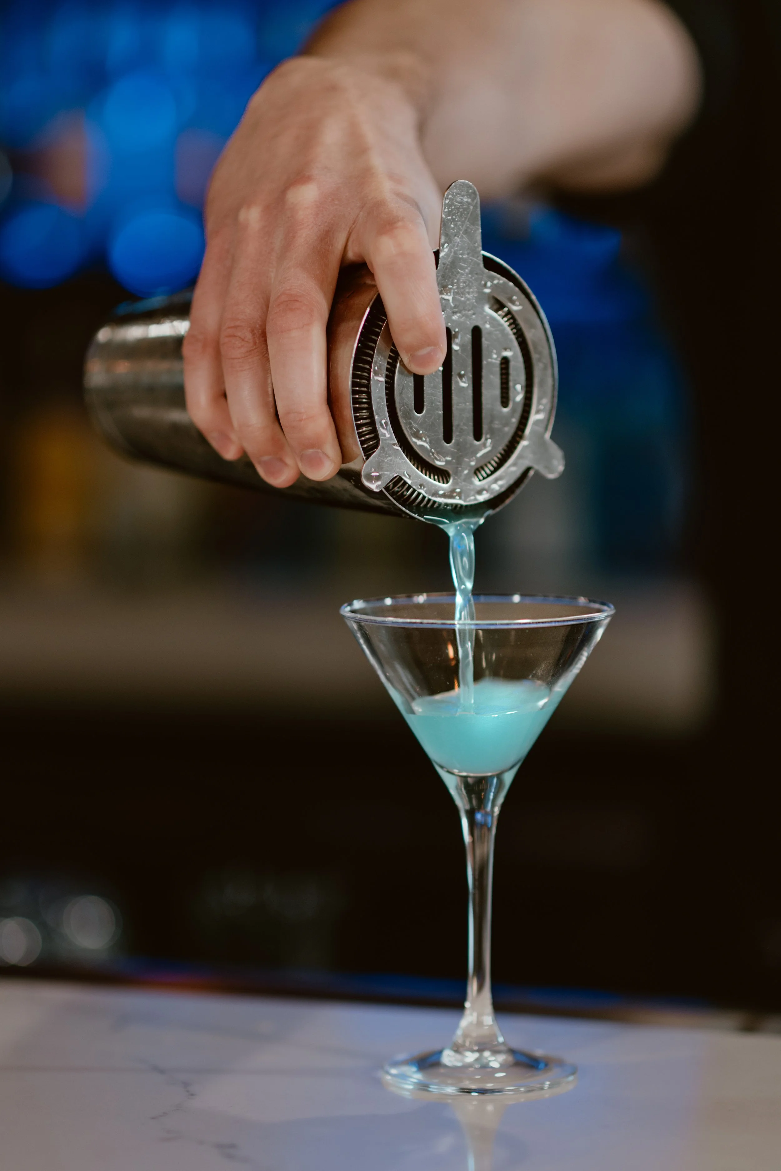 Bartender pouring a blue gin martini from a shaker into a tiny tini glass.