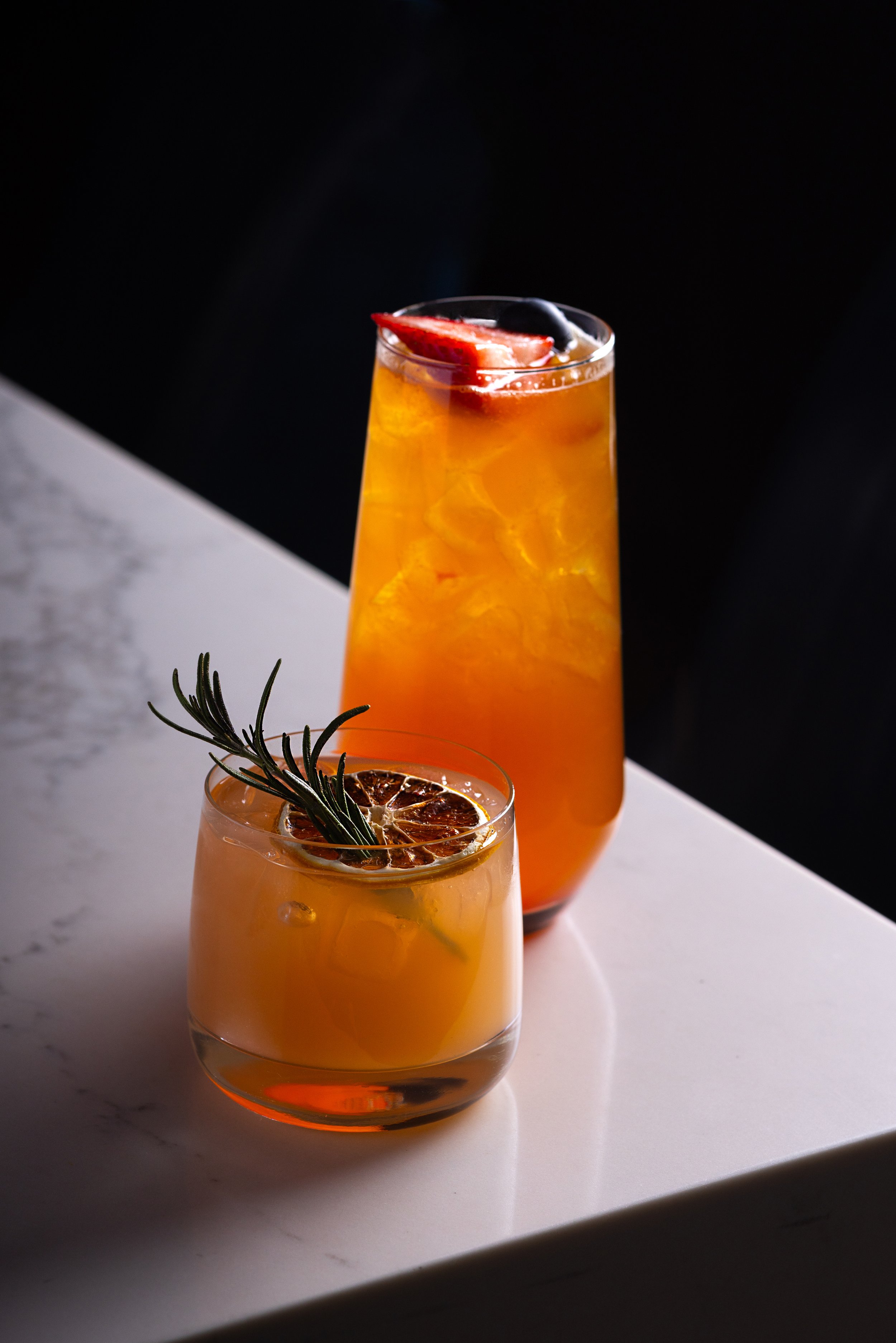 Two gin cocktails on the marble bar top of Southside Shake, one served in a short glass with a sprig of rosemary and a dried citrus slice, and the other in a tall glass with ice and fruit garnishes, against a black background.