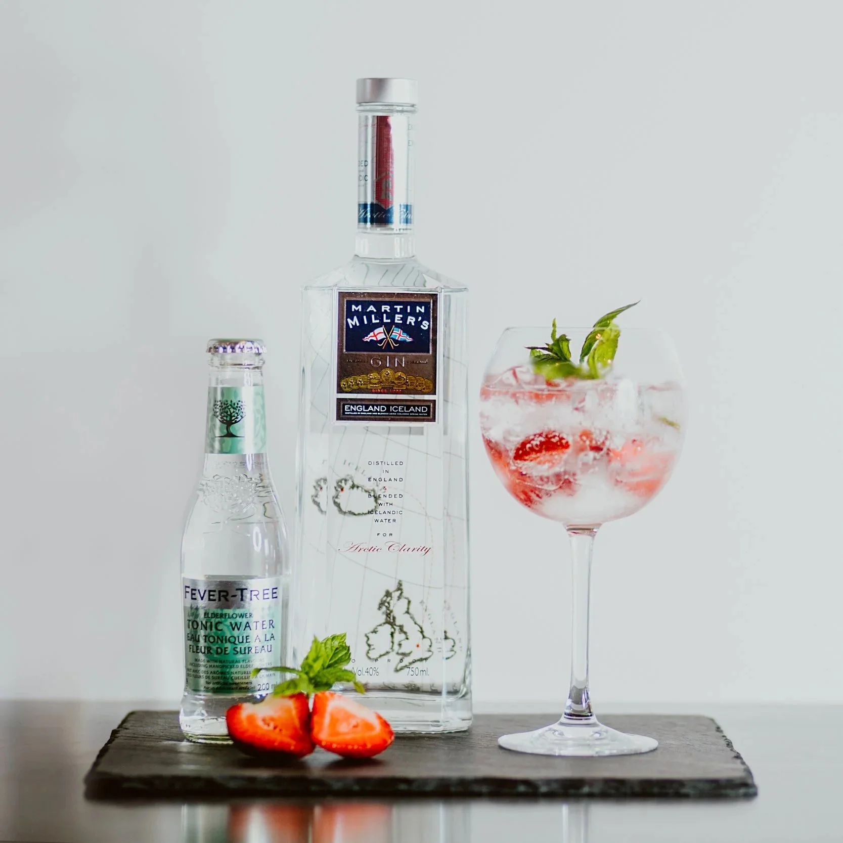 A bottle of Martin Miller's gin, a bottle of Fever-Tree elderflower tonic water, a glass of gin cocktail with strawberries and mint, and a halved strawberry with mint leaves on a dark wooden surface.