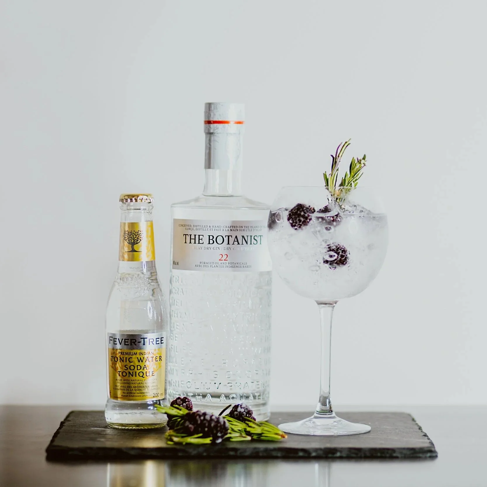 A glass of gin and tonic with blackberries and rosemary garnish, a bottle of Fever-Tree tonic water, and a bottle of Botanist gin on a wooden board.