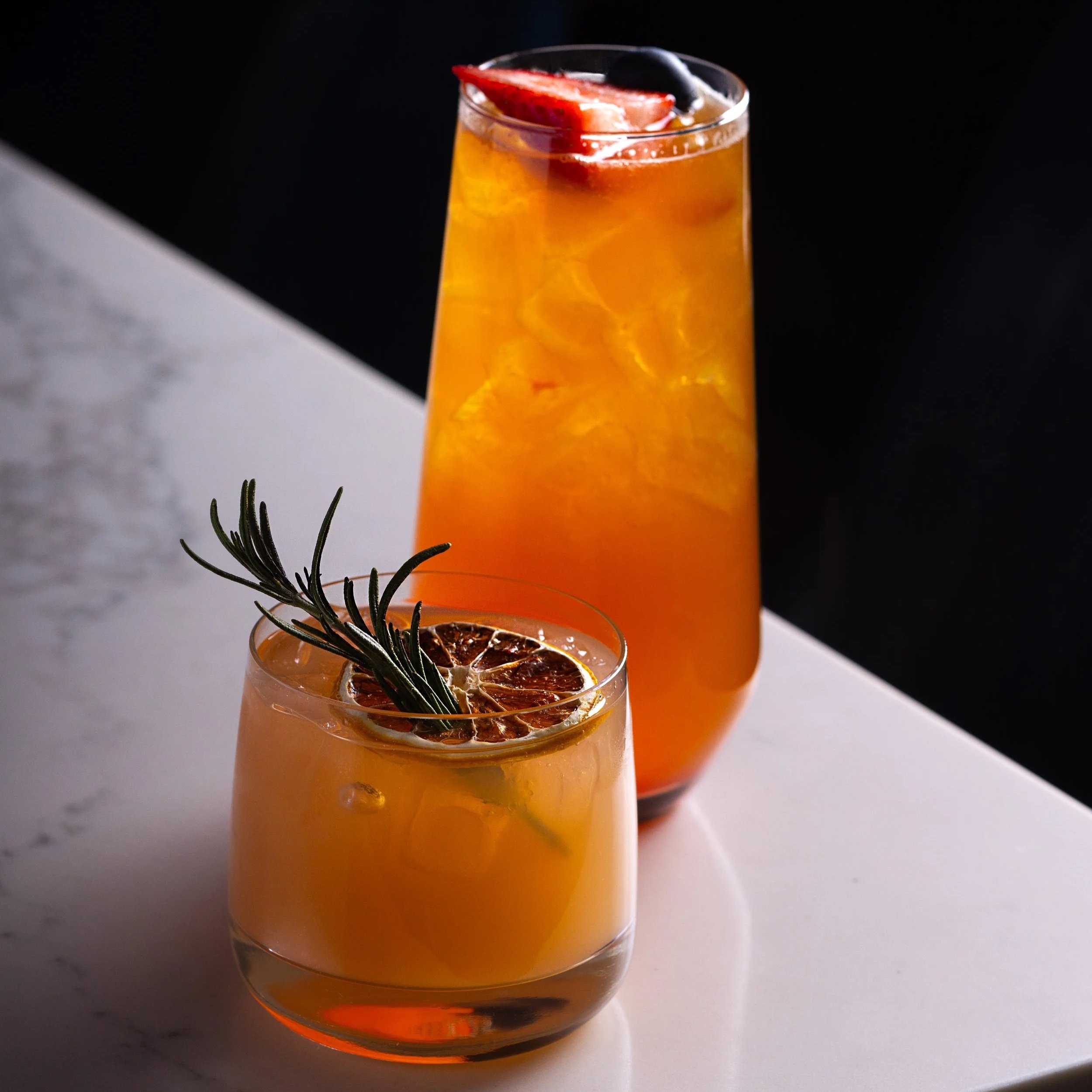 Two gin cocktails on the marble bar top of Southside Shake, one served in a short glass with a sprig of rosemary and a dried citrus slice, and the other in a tall glass with ice and fruit garnishes, against a black background.