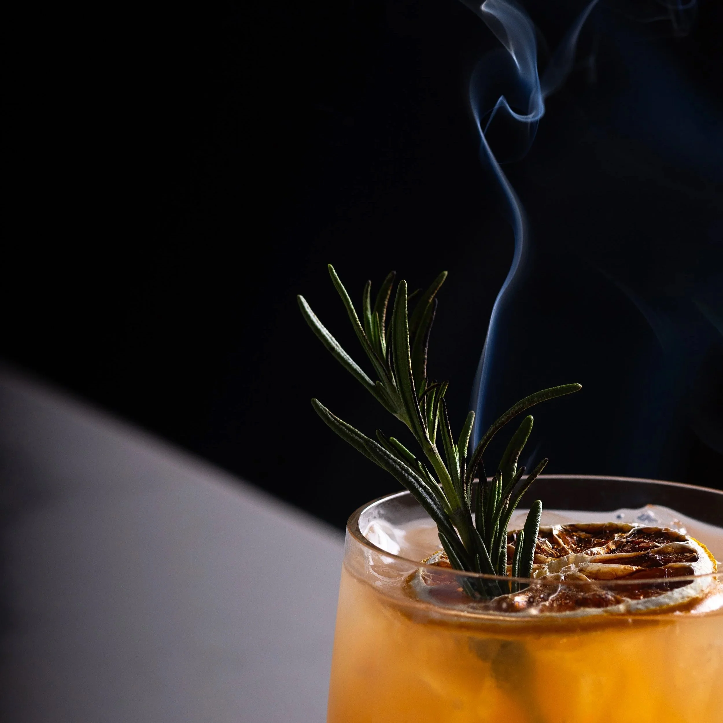 A gin cocktail garnished with a sprig of rosemary and slices of dried citrus, with smoke rising from it, against a dark background at Southside Shake.